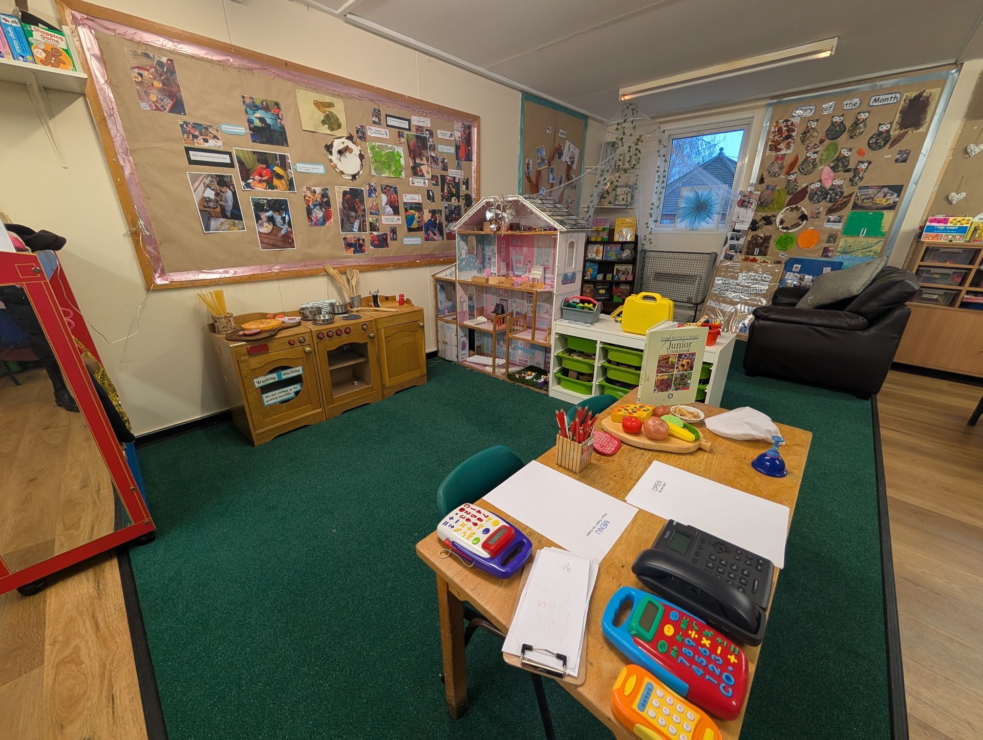 Classroom with bulletin boards, table of supplies, and bookshelves on a green carpet.