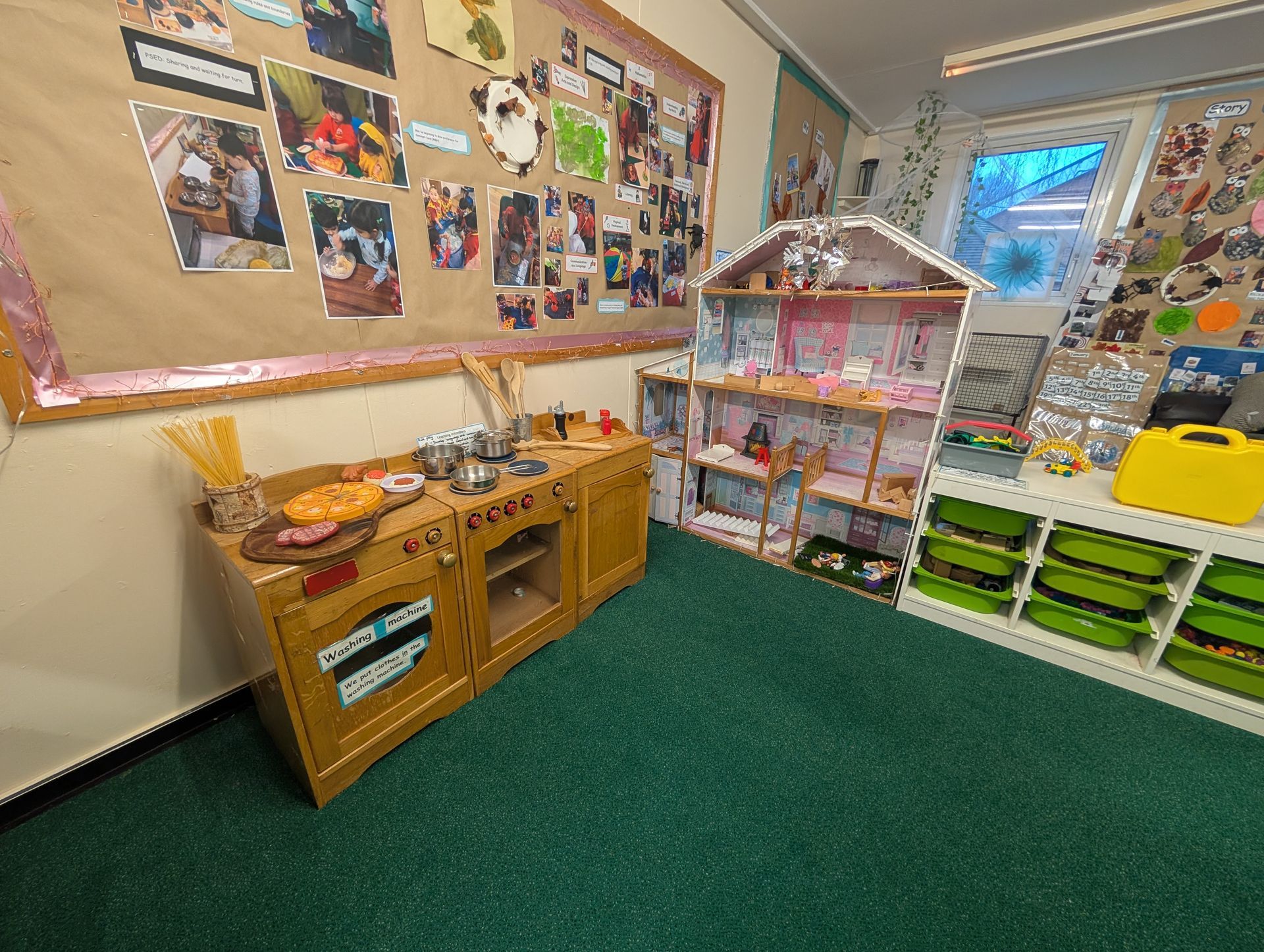 Childcare playroom with toy kitchen, dollhouse, bulletin boards, and green carpet