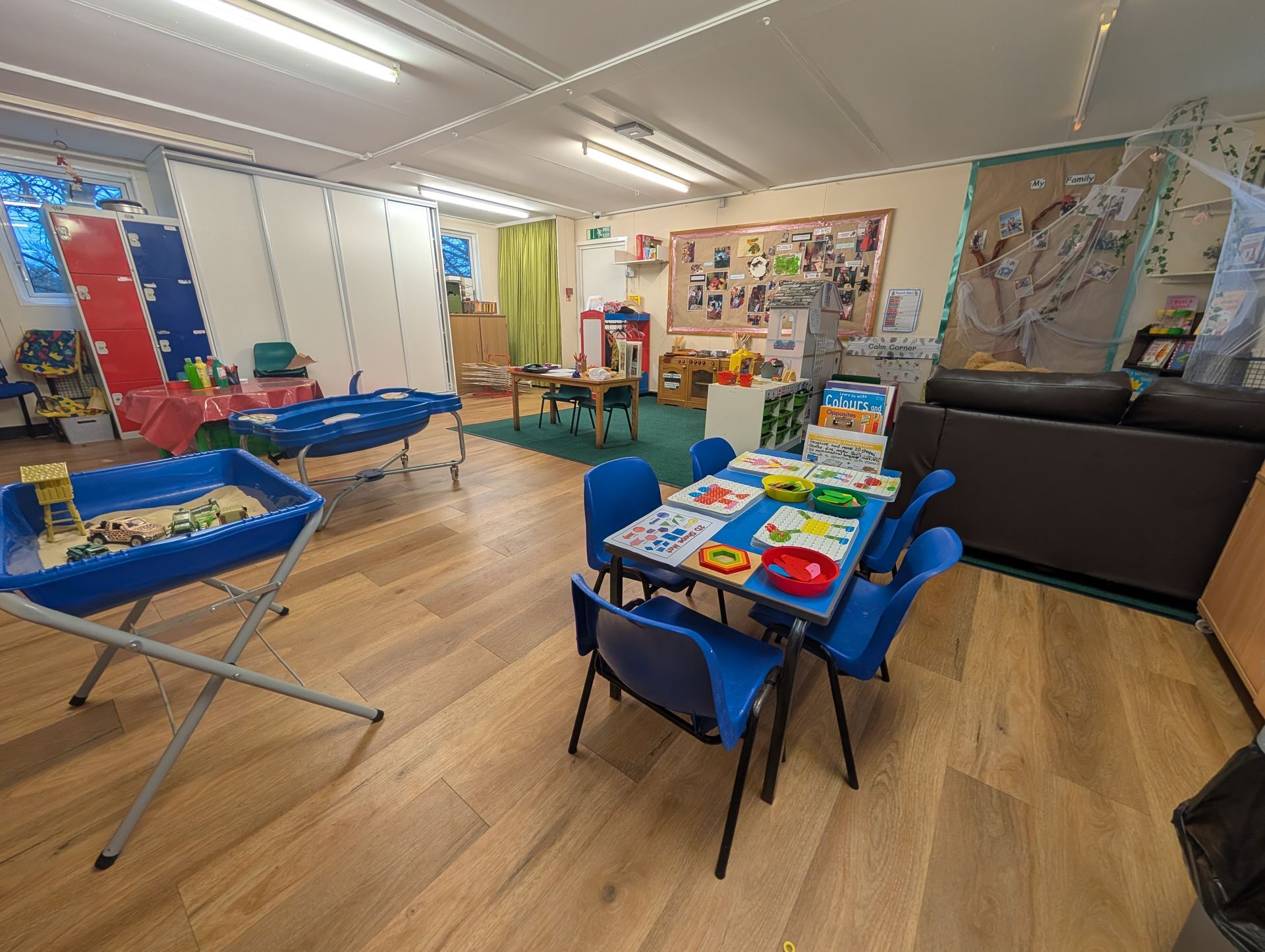 Bright classroom with toy tables, blue chairs, rugs, and colorful wall displays.