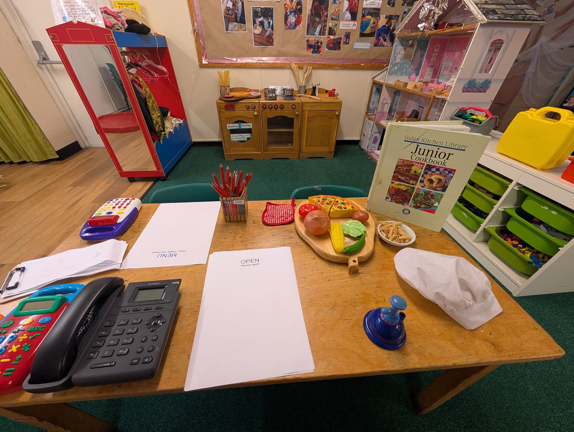 Classroom table with papers, crayons, toys, and a toy cash register in a colorful play area