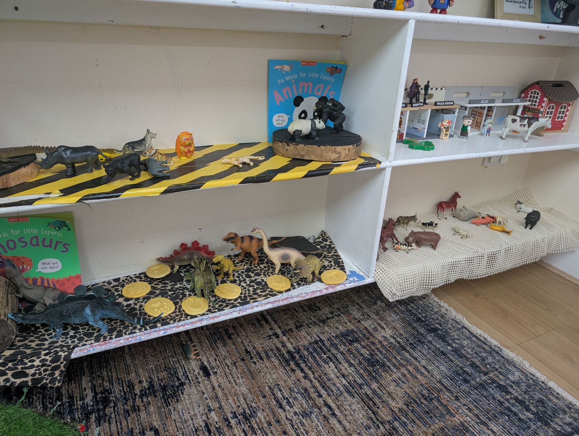 Toy shelves with dinosaur playsets, animal figures, and books on white cubbies over a rug.