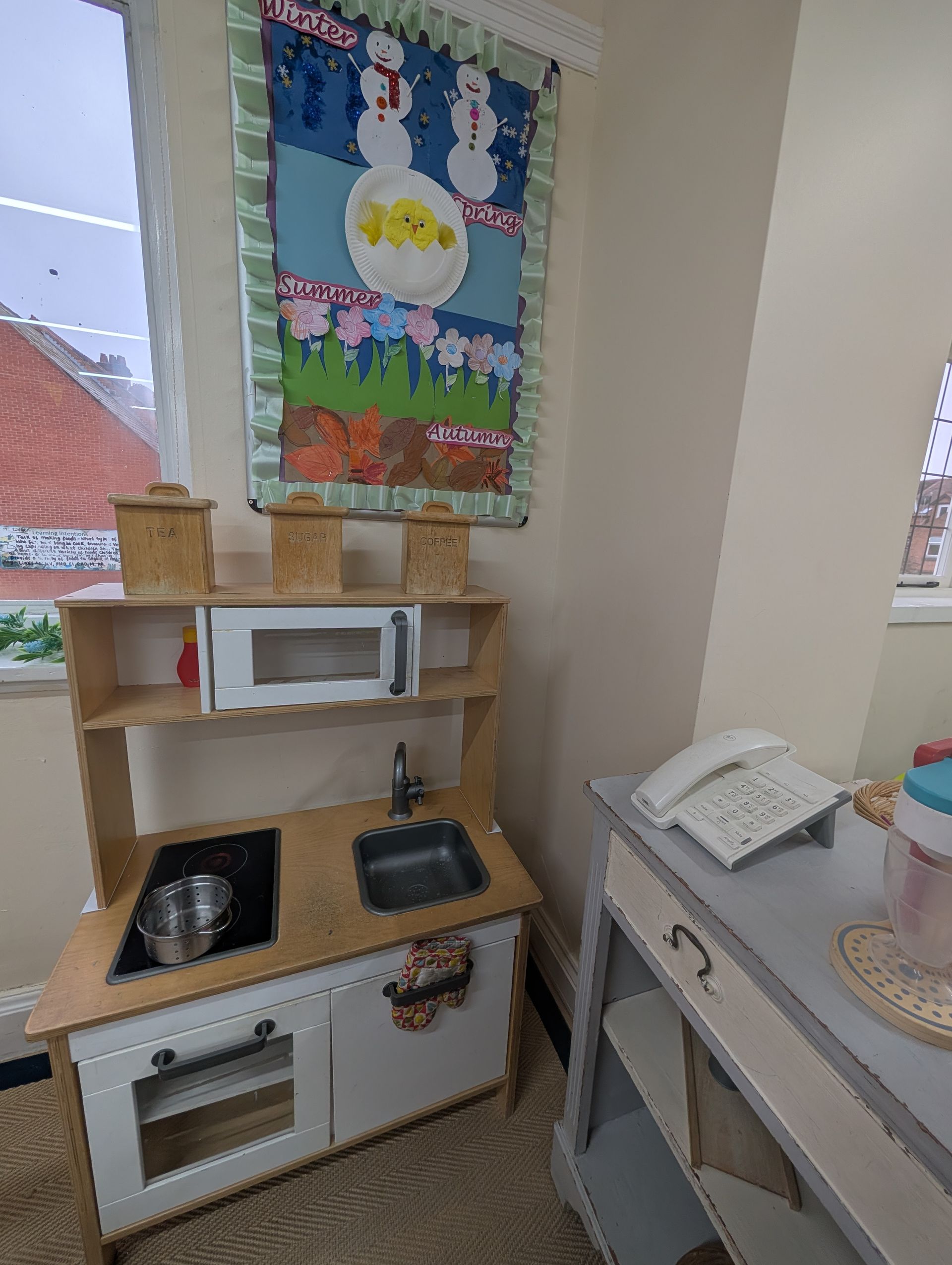 Small play kitchen with toy microwave, stove, and phone beside a window and colorful wall hanging