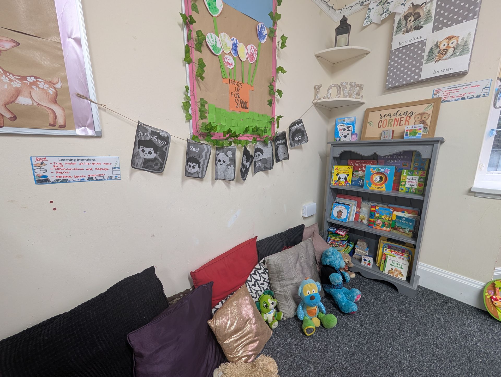 Cozy corner with colorful pillows, stuffed animals, wall art, and a toy bookshelf in a child’s room.