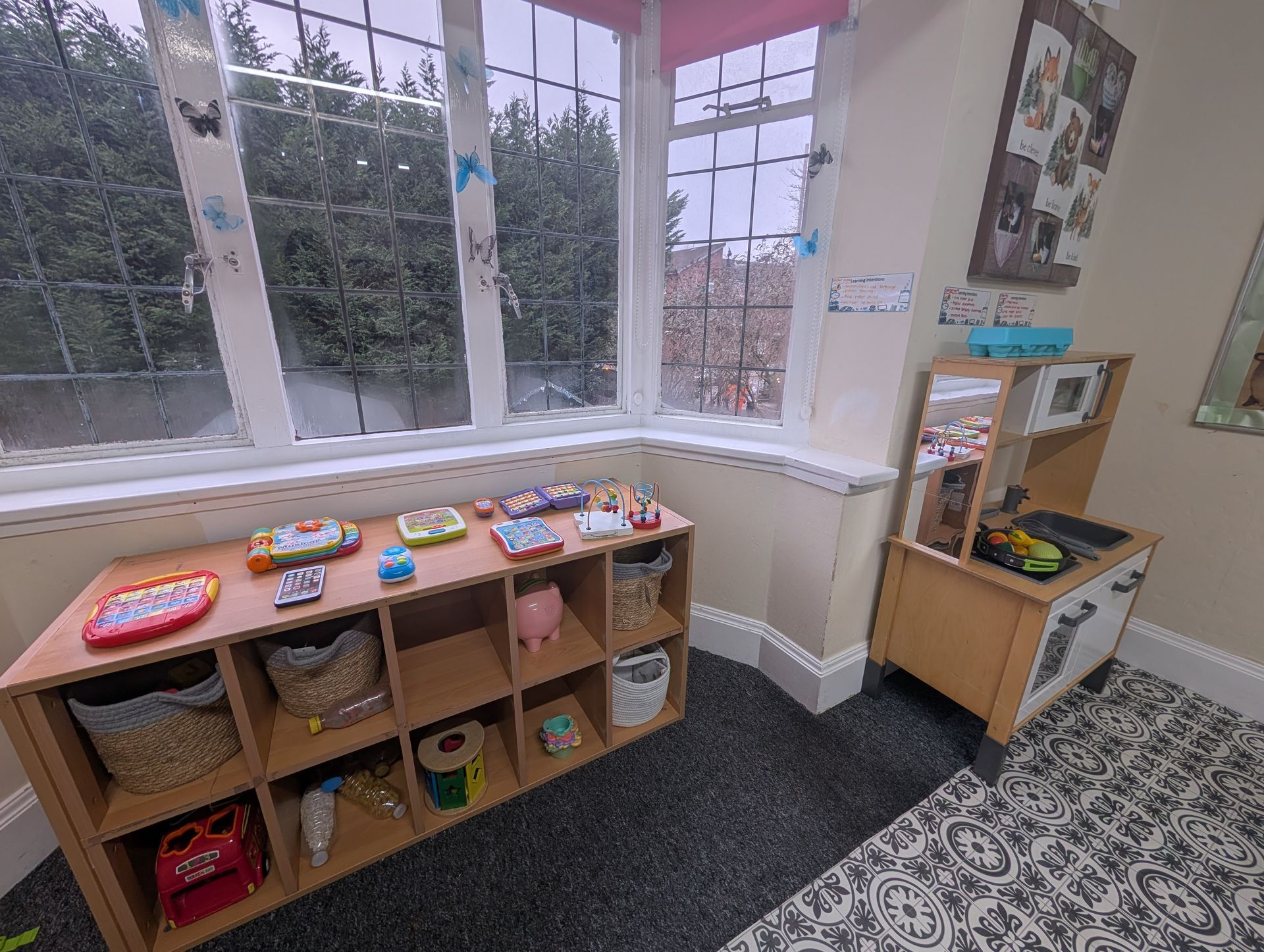 Childcare play area with toys on low shelves by large windows, patterned carpet, and a small wooden play kitchen