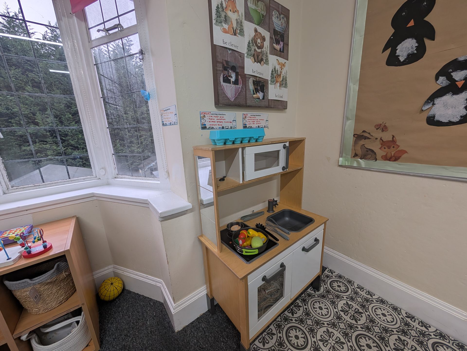 Children’s play kitchen in a bright corner by a window, with patterned rug and wall art