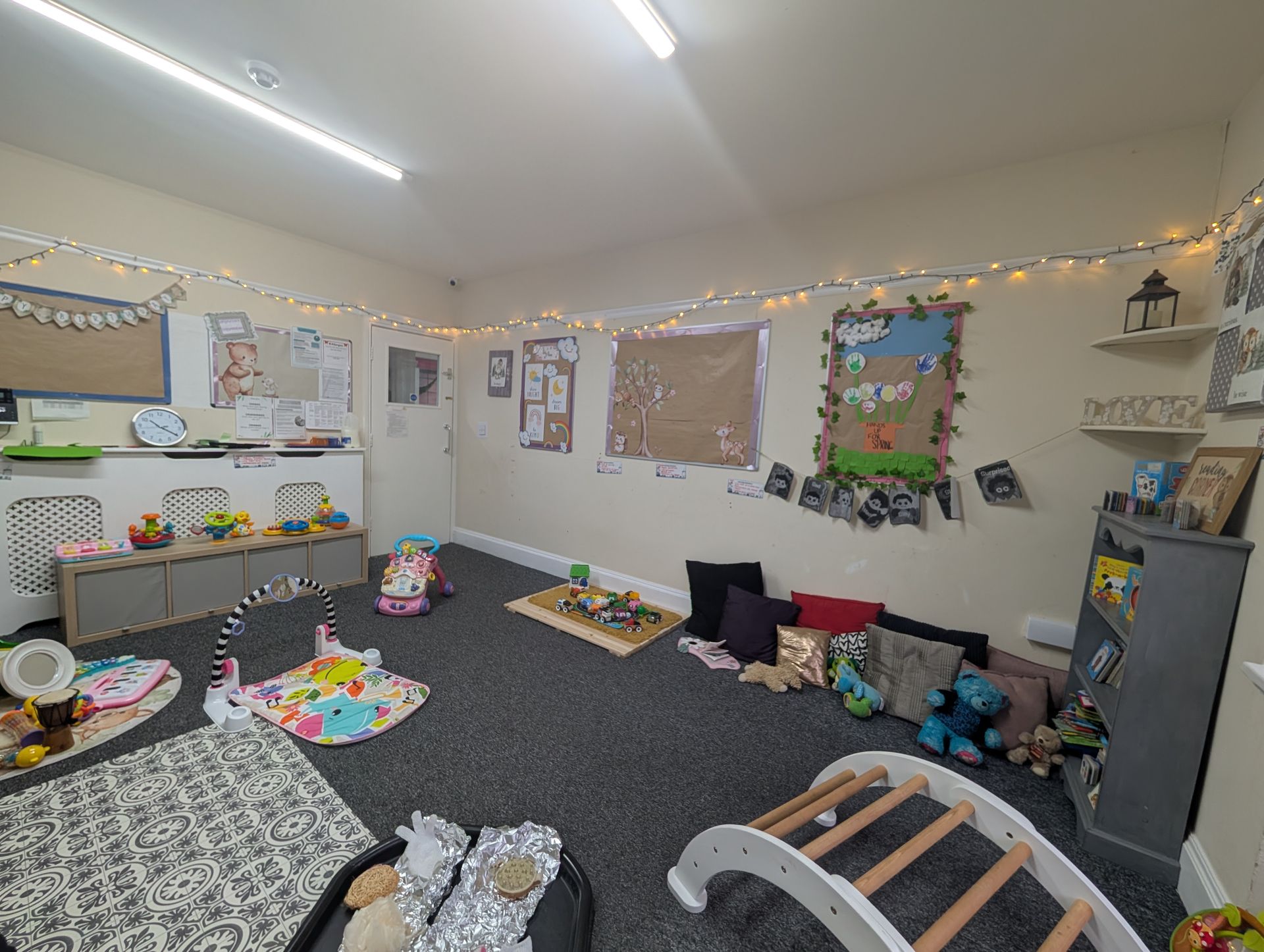Bright classroom corner with rugs, toys, bulletin boards, and string lights on the walls.