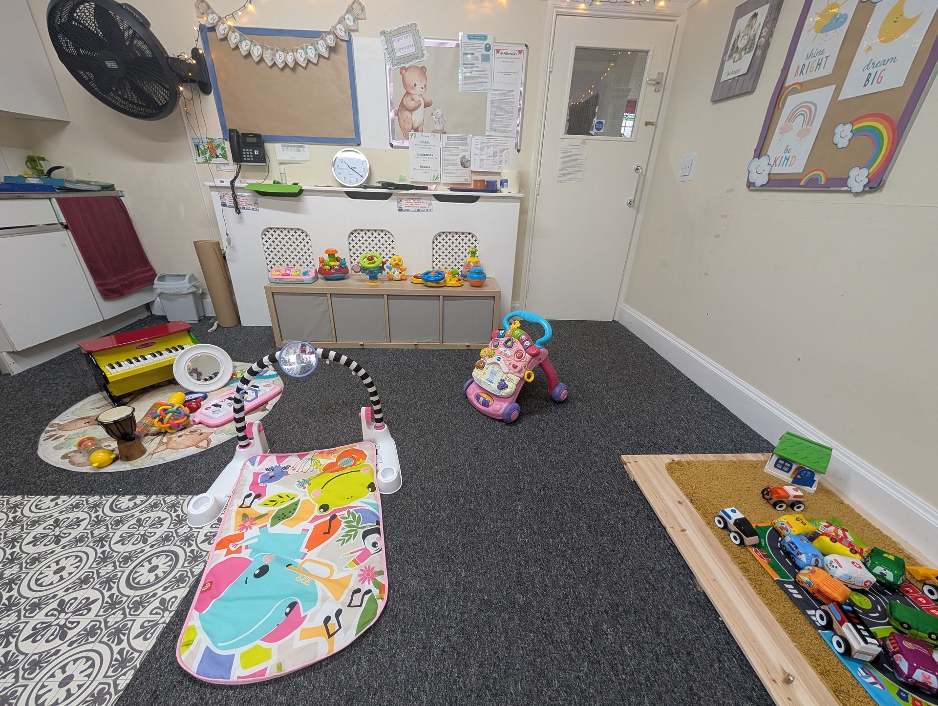 Playroom with scattered toys, colorful mats, a storage shelf, and wall decorations on a carpeted floor.