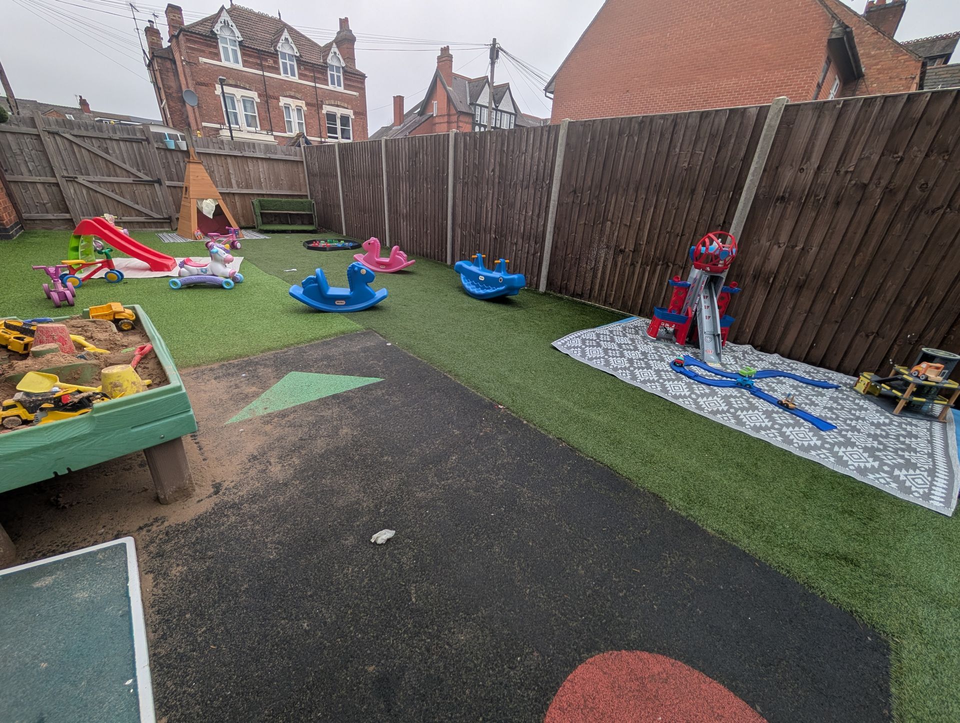 Backyard playground with toys, a small slide, and a Spider-Man figure on a patio mat near a wooden fence
