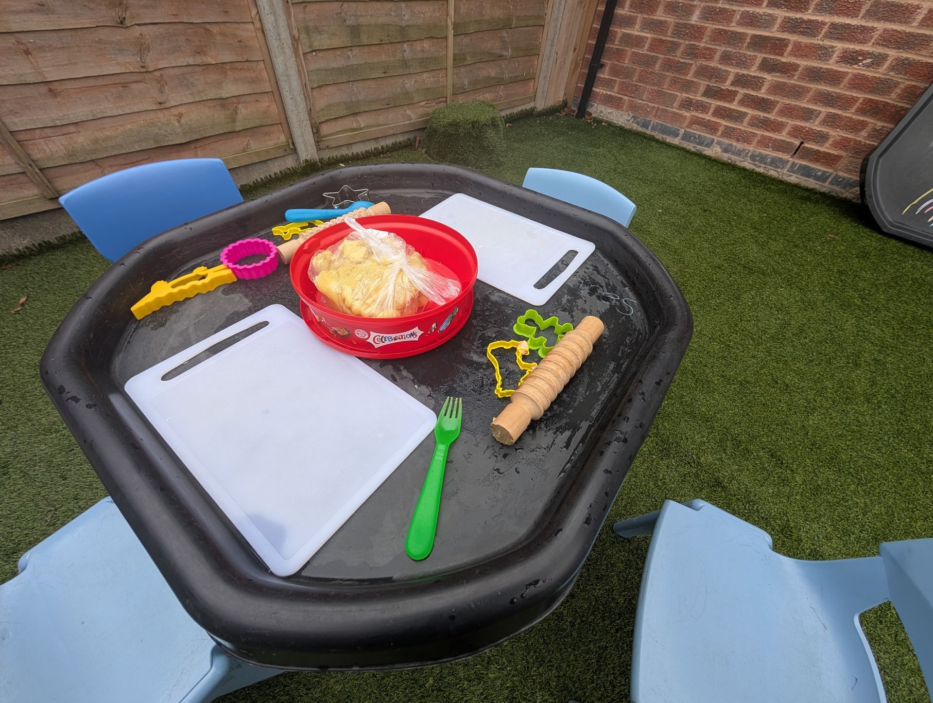 Child’s outdoor play table with toy kitchen items, bowls, utensils, and plastic food on a patio.