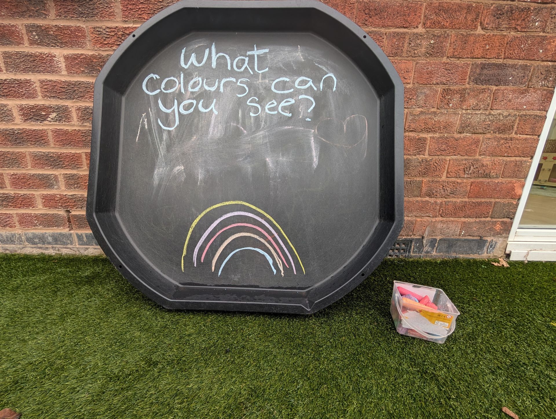 Chalkboard asks “what colours can you see?” with a rainbow drawing beside a brick wall and grass