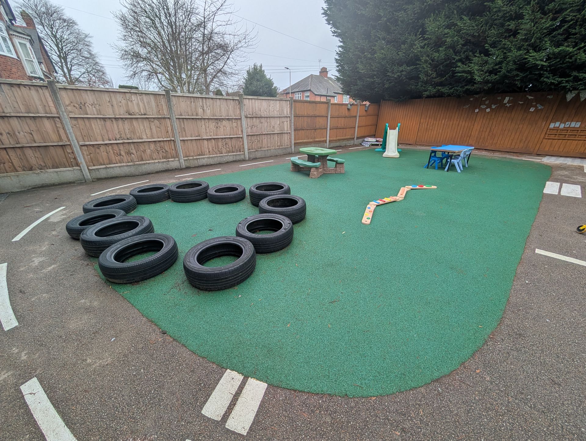 Outdoor playground area with a large tire obstacle course on green turf and a wooden fence around it.