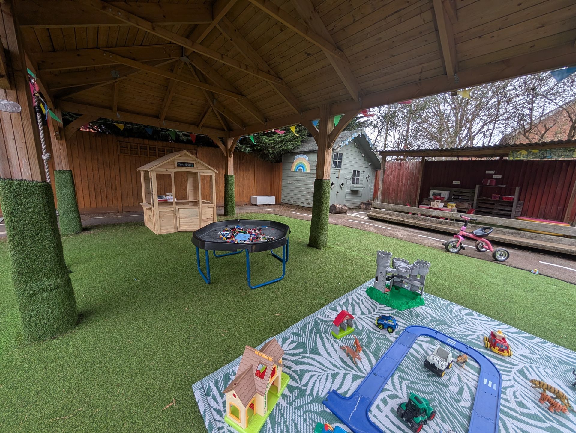 Covered play area with artificial grass, toy play mat, wooden playhouse, and a small trampoline outdoors
