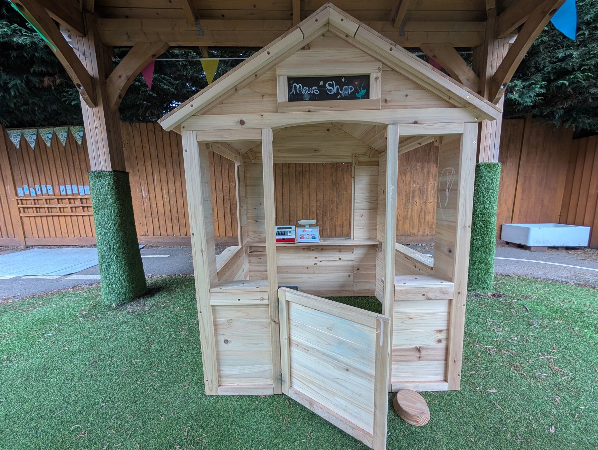 Small wooden playhouse with an open door under a covered outdoor area, on artificial grass.