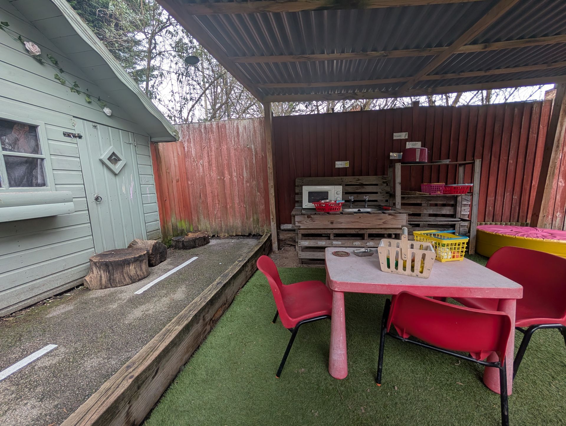 Covered backyard patio with pink table, red chairs, grill, and wooden fence.