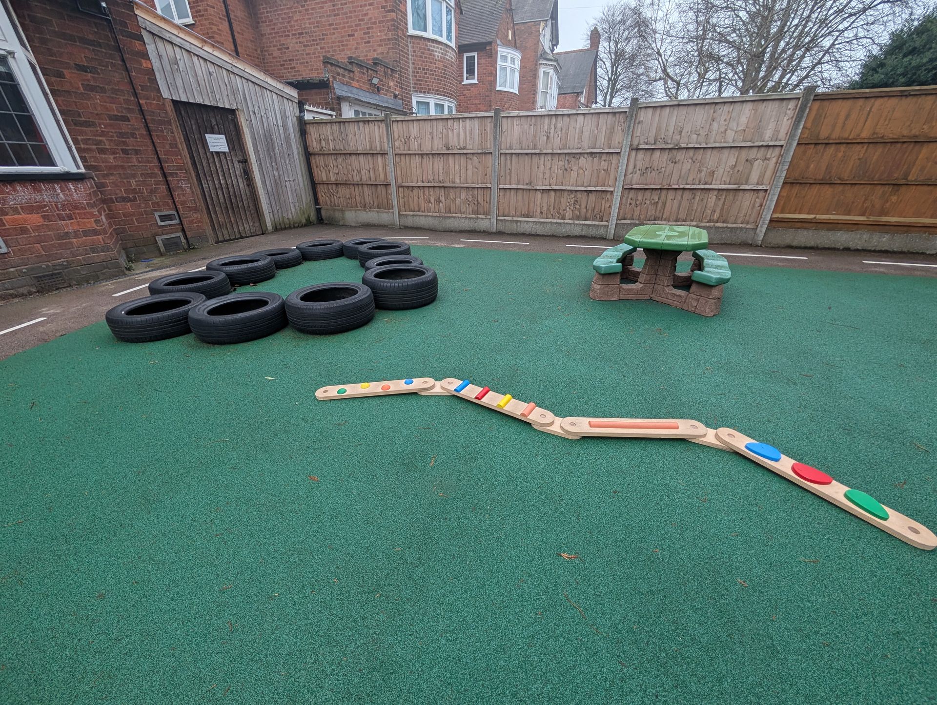 Outdoor play area with green turf, stacked tires, a small picnic table, and a colorful wooden path.