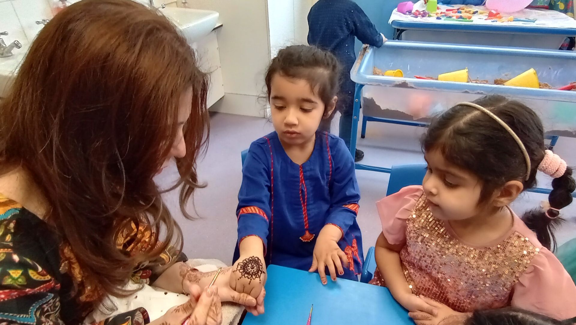 Two children and an adult sit at a table, interacting with small objects in a classroom setting.