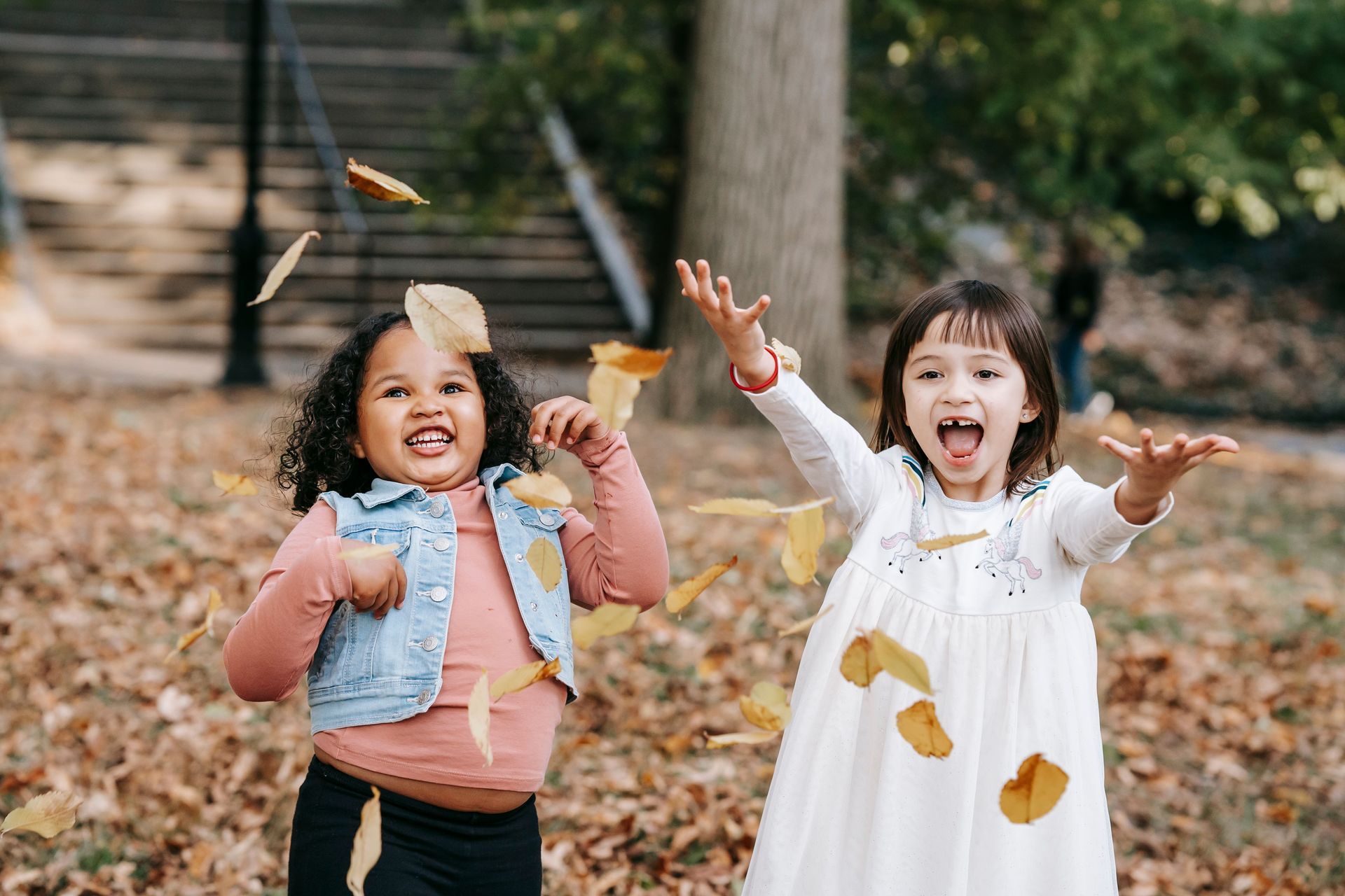 Two children playing in autumn leaves outdoors, smiling with arms raised near a wooden fence.