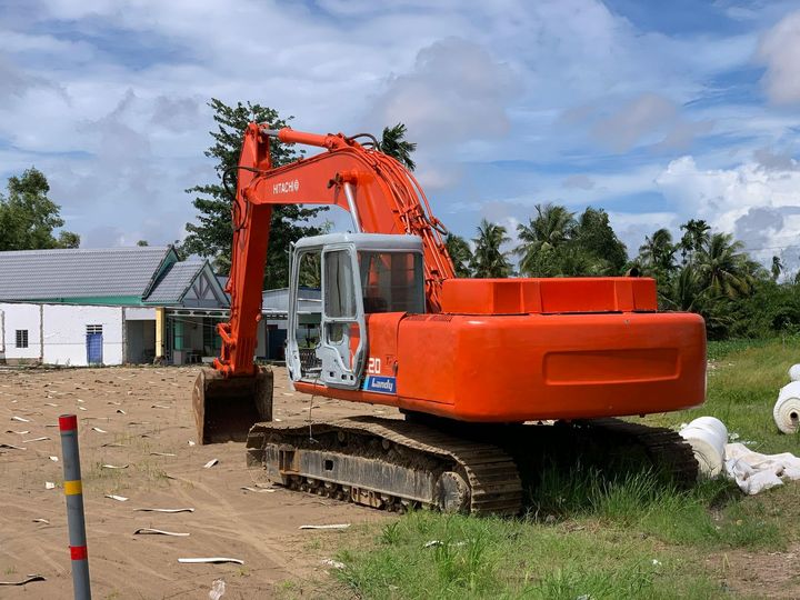 An orange excavator sits on a dirt lot in front of a white building under a blue sky with scattered clouds.