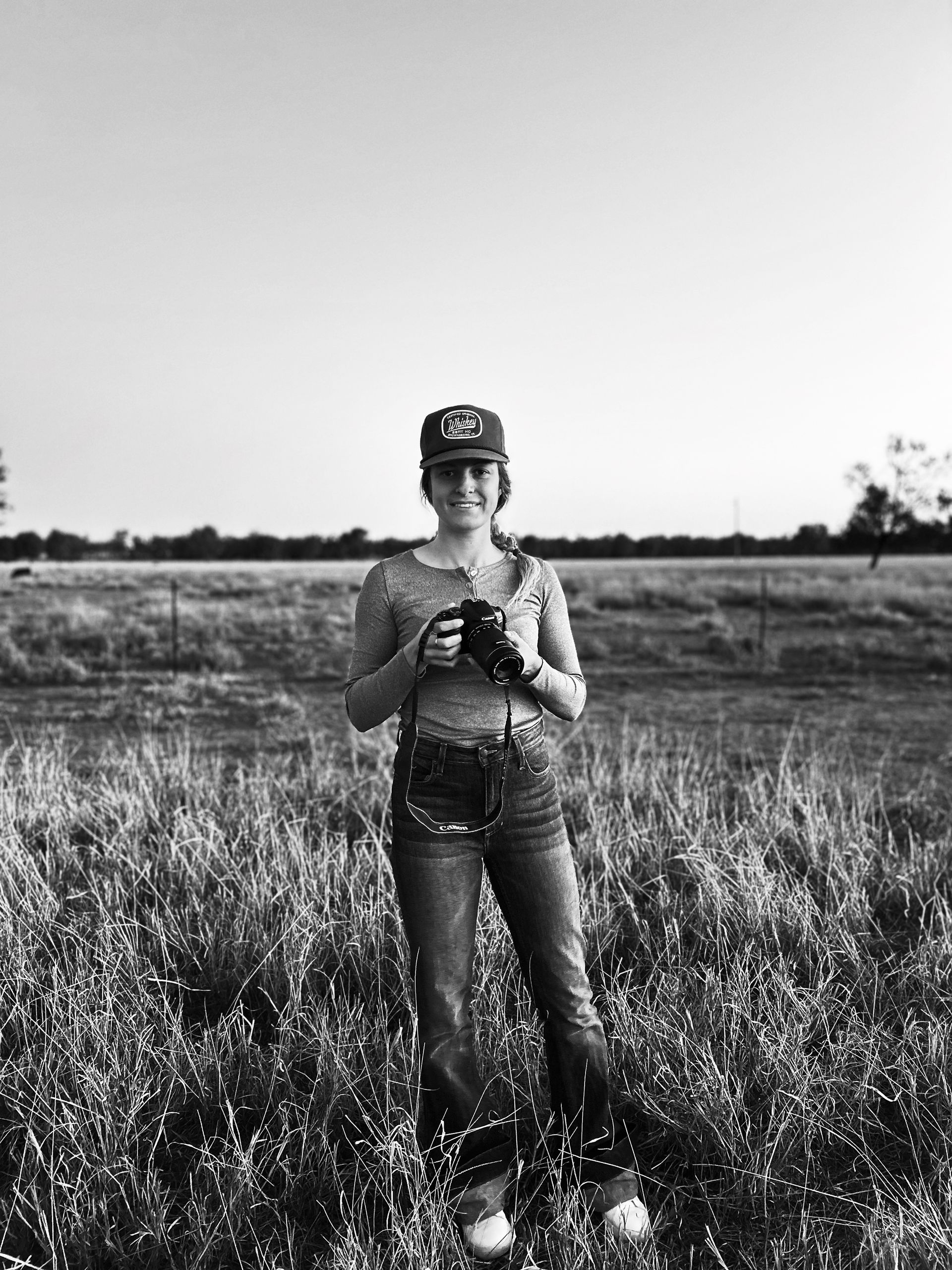 Person standing in a field, holding a camera, with tall grass and open sky behind them.