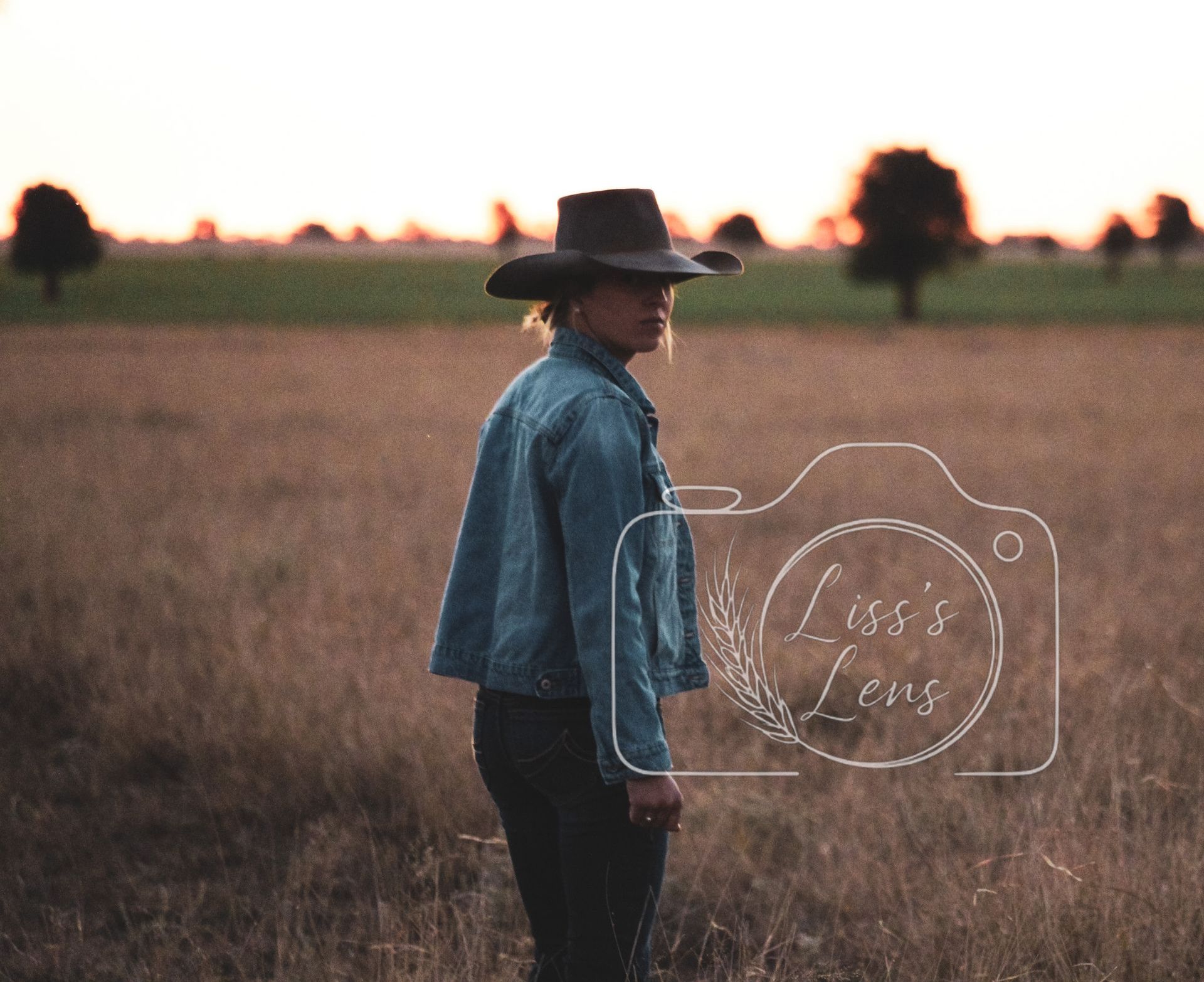 Person in a denim jacket and cowboy hat standing in a field at sunset.