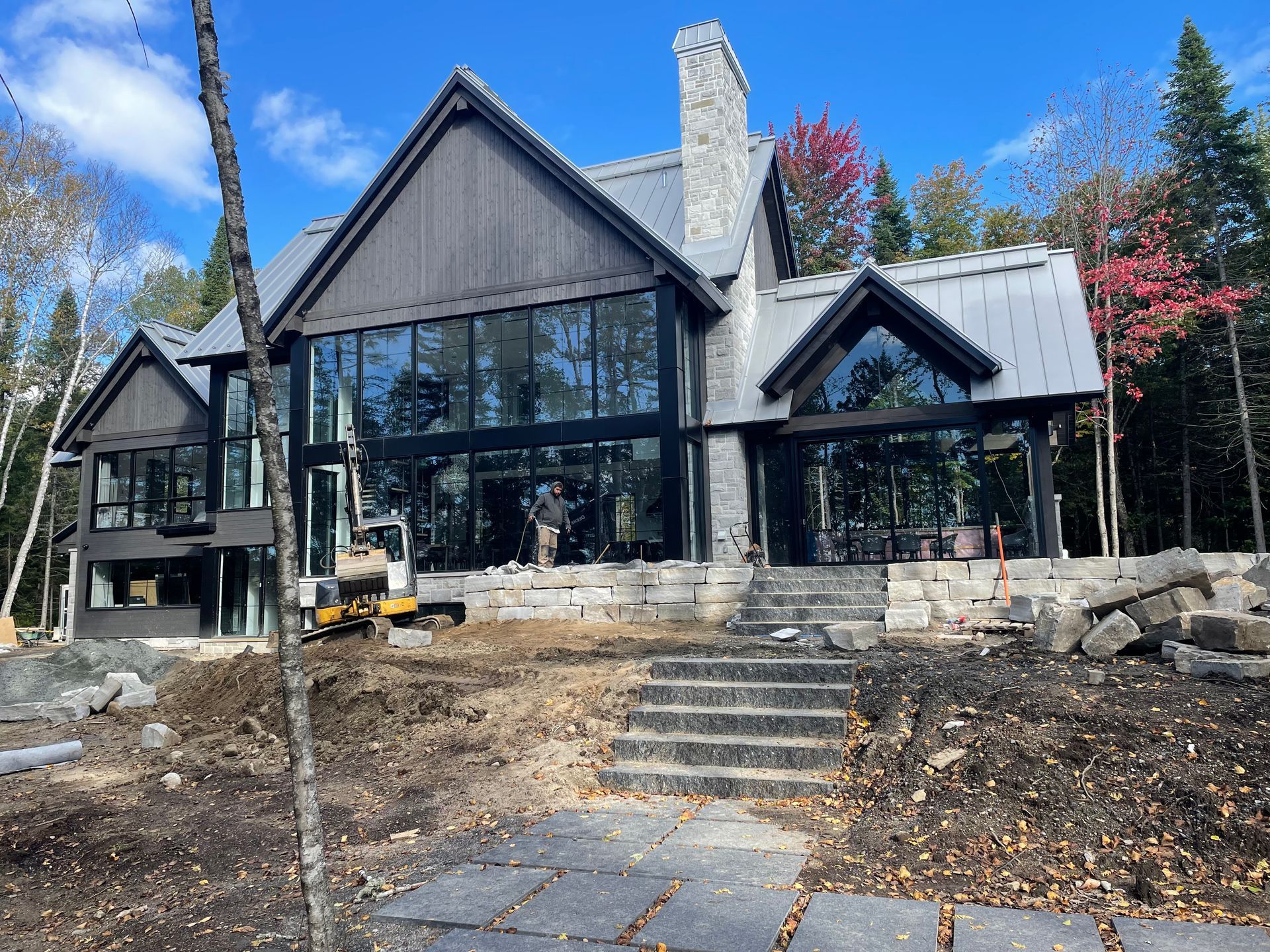 Maison moderne en construction avec de grandes baies vitrées et un escalier en pierre. Toiture et façade grises, avec une petite excavatrice.