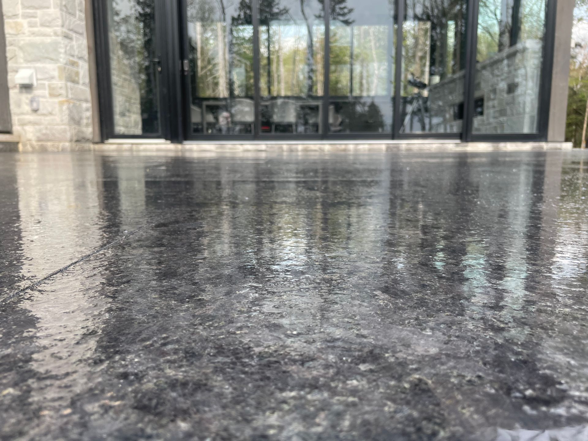 Gros plan d'un patio en béton gris foncé et humide reflétant une porte vitrée et des arbres.
