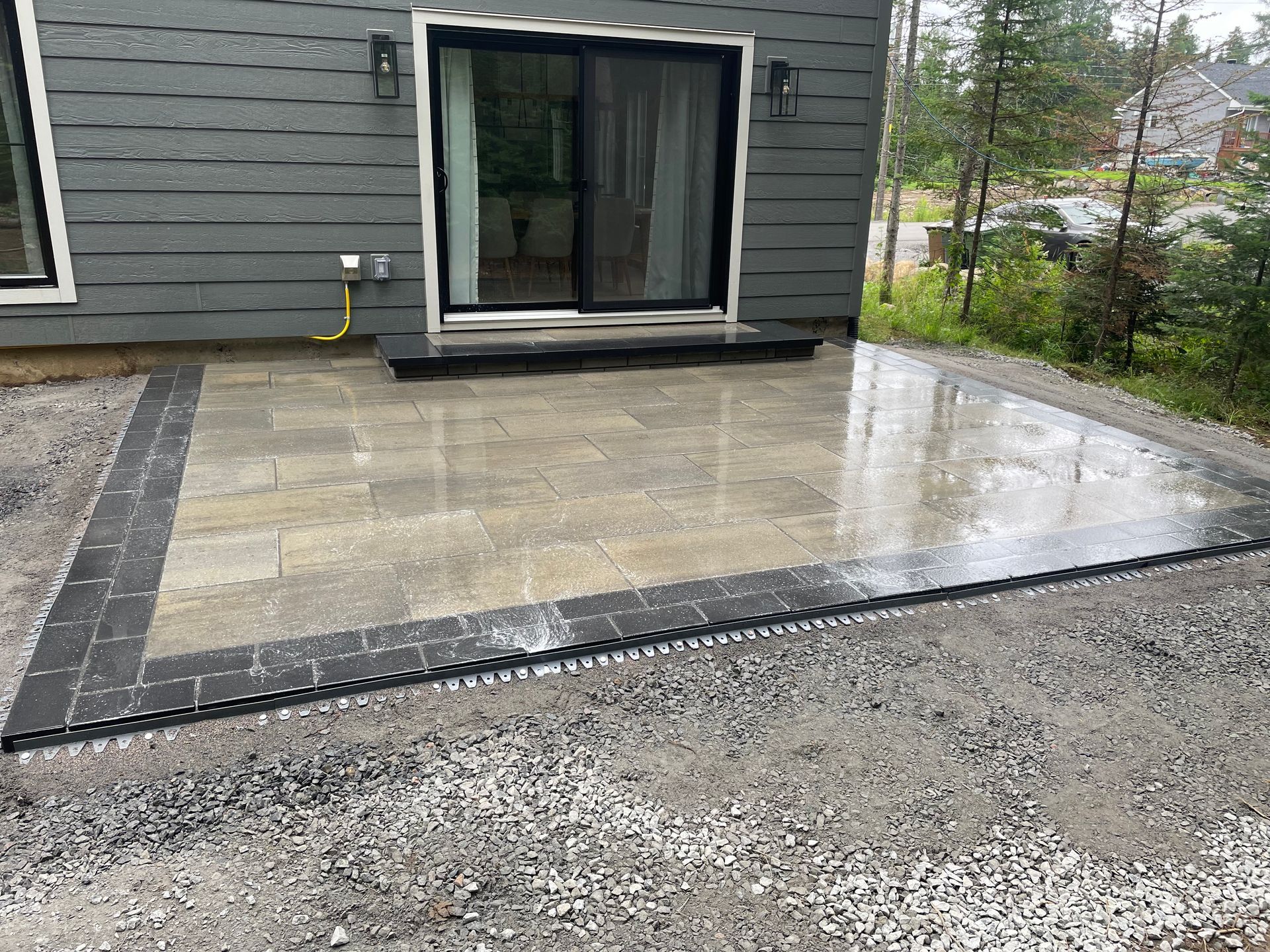 Terrasse en briques grises à côté d'une maison avec portes coulissantes. La terrasse est humide et bordée de noir.