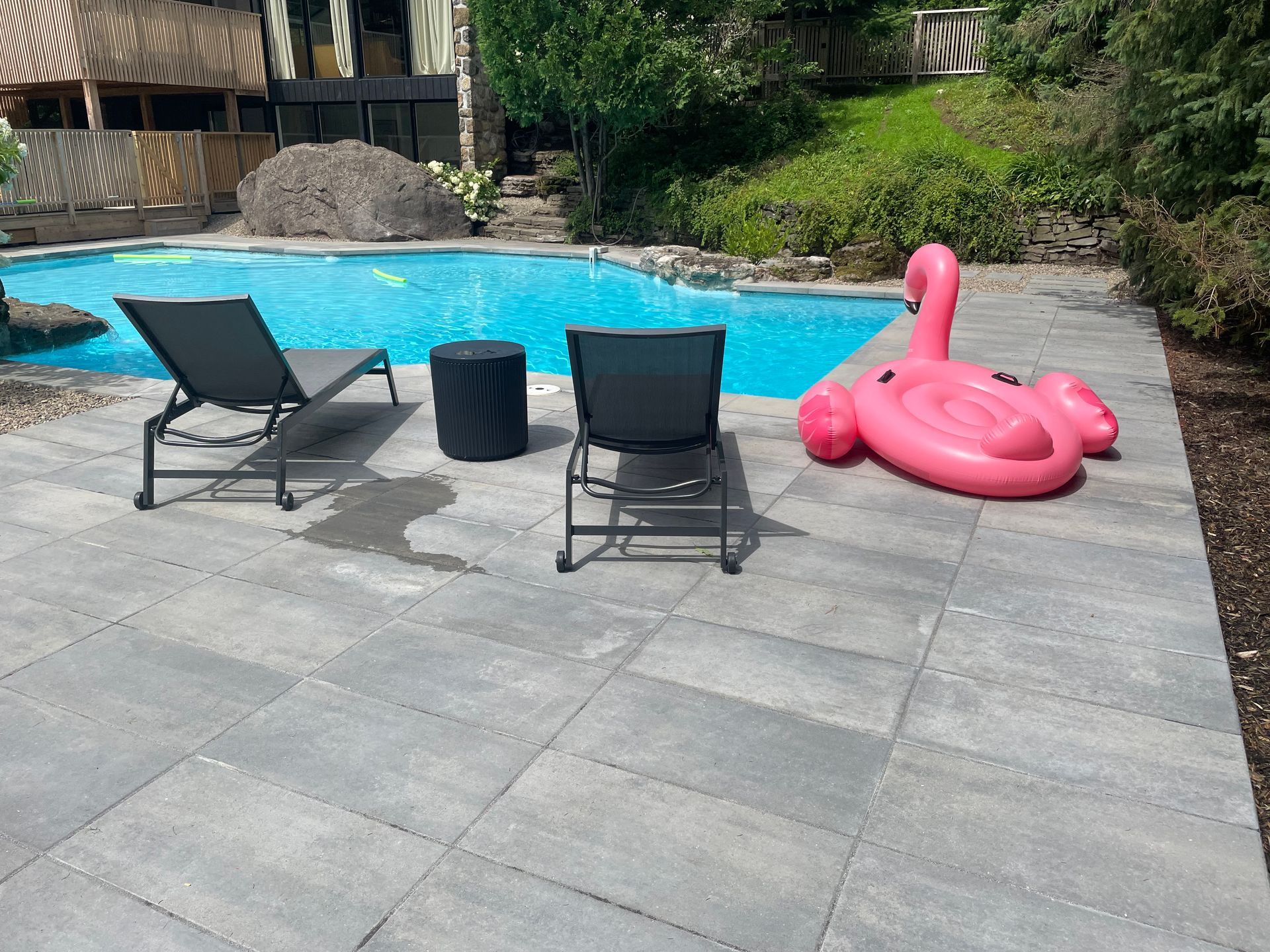 Scène au bord de la piscine avec une piscine bleue, deux chaises longues, une table noire et un flotteur de flamant rose sur un patio gris.