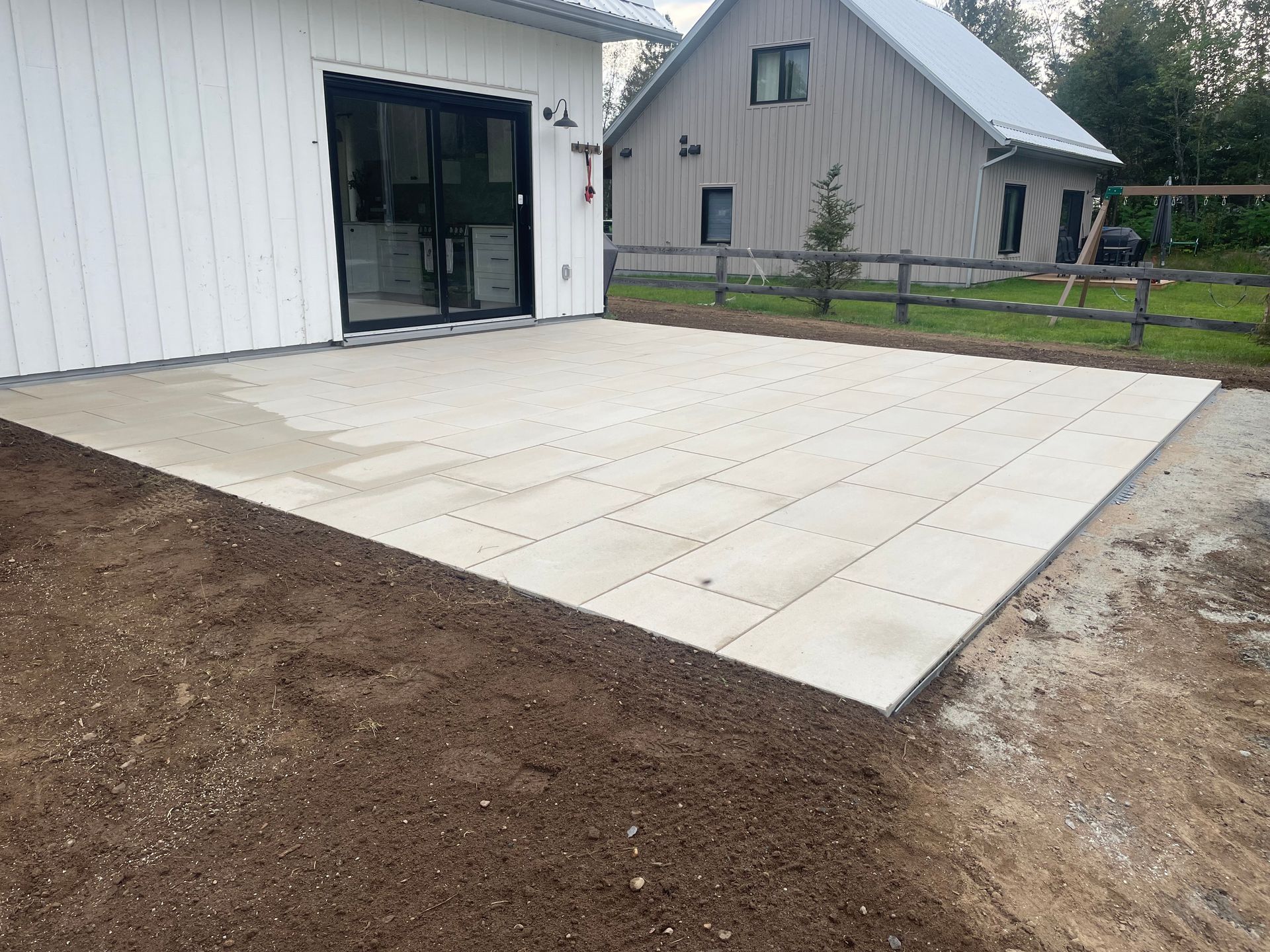 Un patio en béton nouvellement construit à côté d'un bâtiment blanc, avec de la terre environnante et une deuxième maison en arrière-plan.