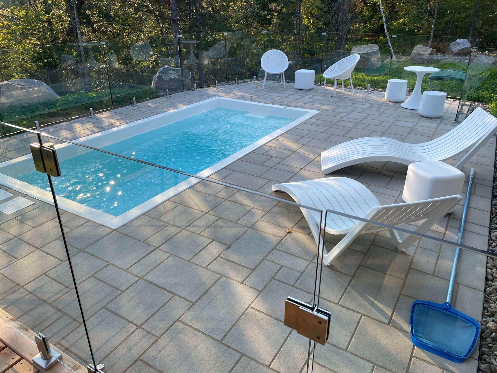 Une petite piscine rectangulaire entourée d'un patio en pierre avec des chaises longues blanches et une clôture en verre.
