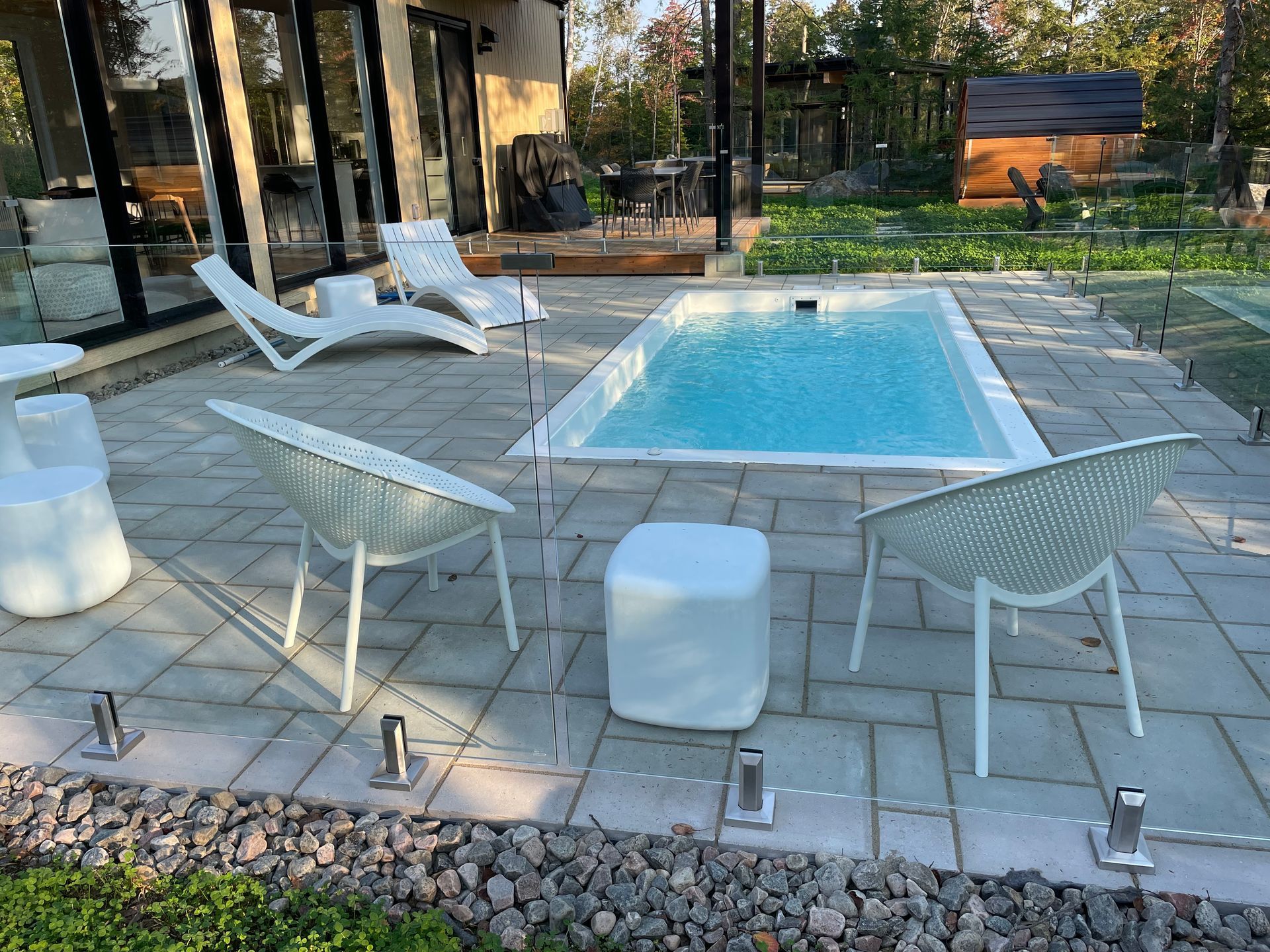 Un jardin moderne avec une petite piscine rectangulaire, du mobilier de jardin et une clôture en verre. Des chaises et des transats blancs entourent la piscine sur un patio en pierre.