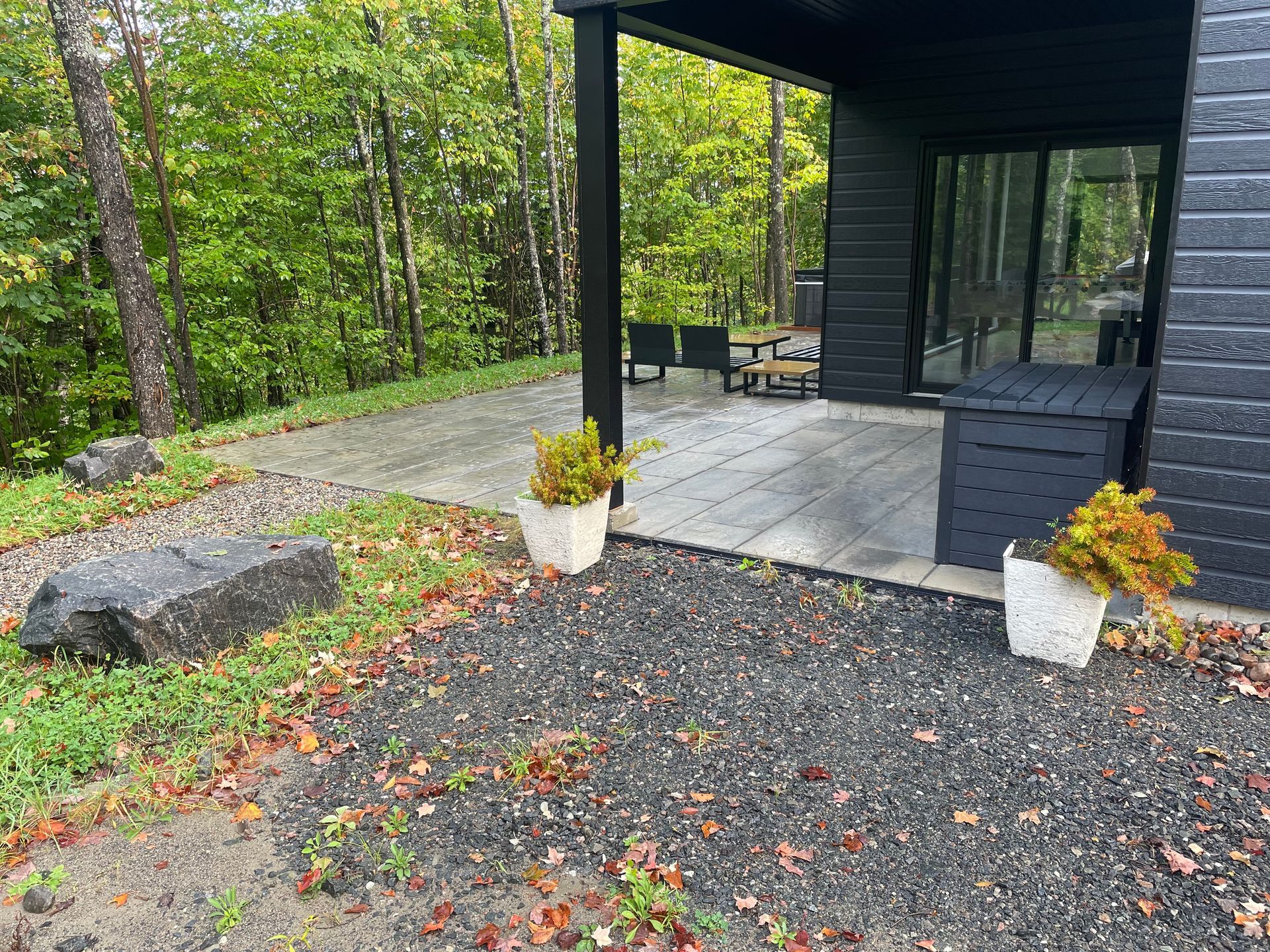 Un patio moderne gris foncé avec du gravier, de grosses pierres et des plantes en pot. Il jouxte une maison sombre dotée de grandes baies vitrées coulissantes, avec une forêt en arrière-plan.