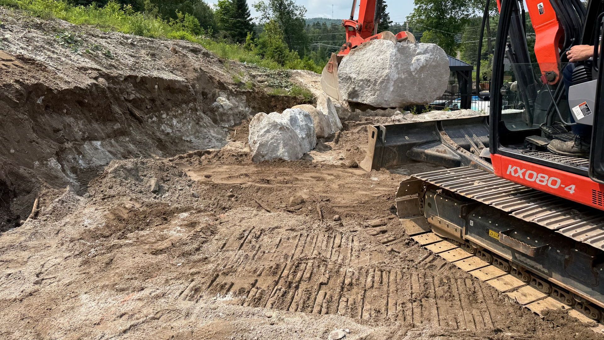 Une excavatrice orange déplace de gros rochers sur un terrain en terre. La machine est posée sur une surface de terre brune.