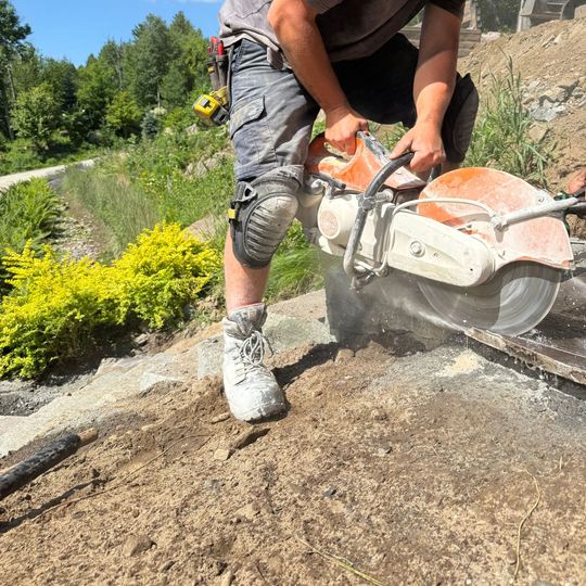 Un homme utilise une scie à béton à l'extérieur pour découper une surface grise. Il porte un short gris, des genouillères et des bottes de travail ; l'eau gicle.
