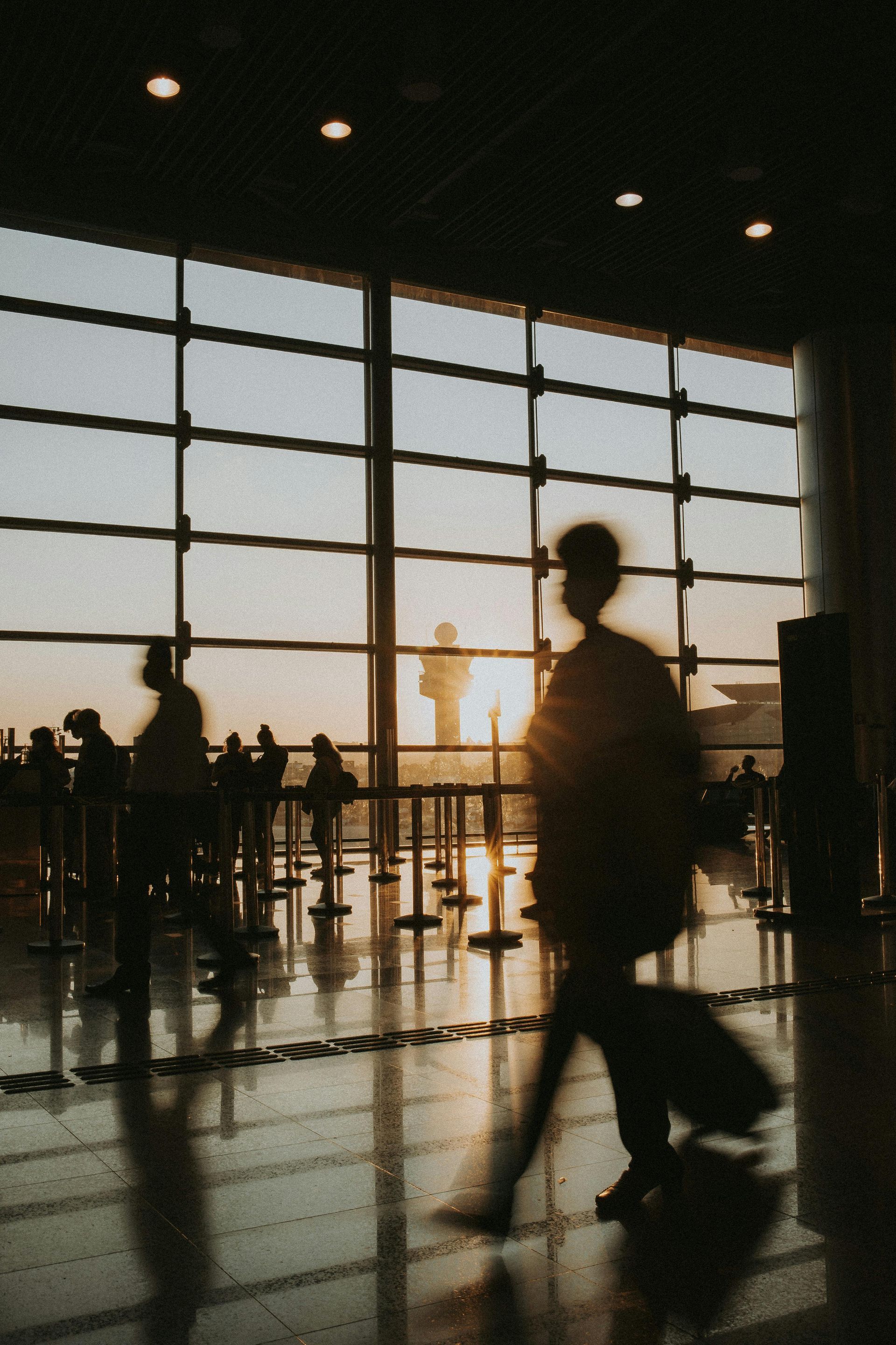 Silhouetted travelers walk through an airport terminal against a backdrop of a bright sunset through large windows.