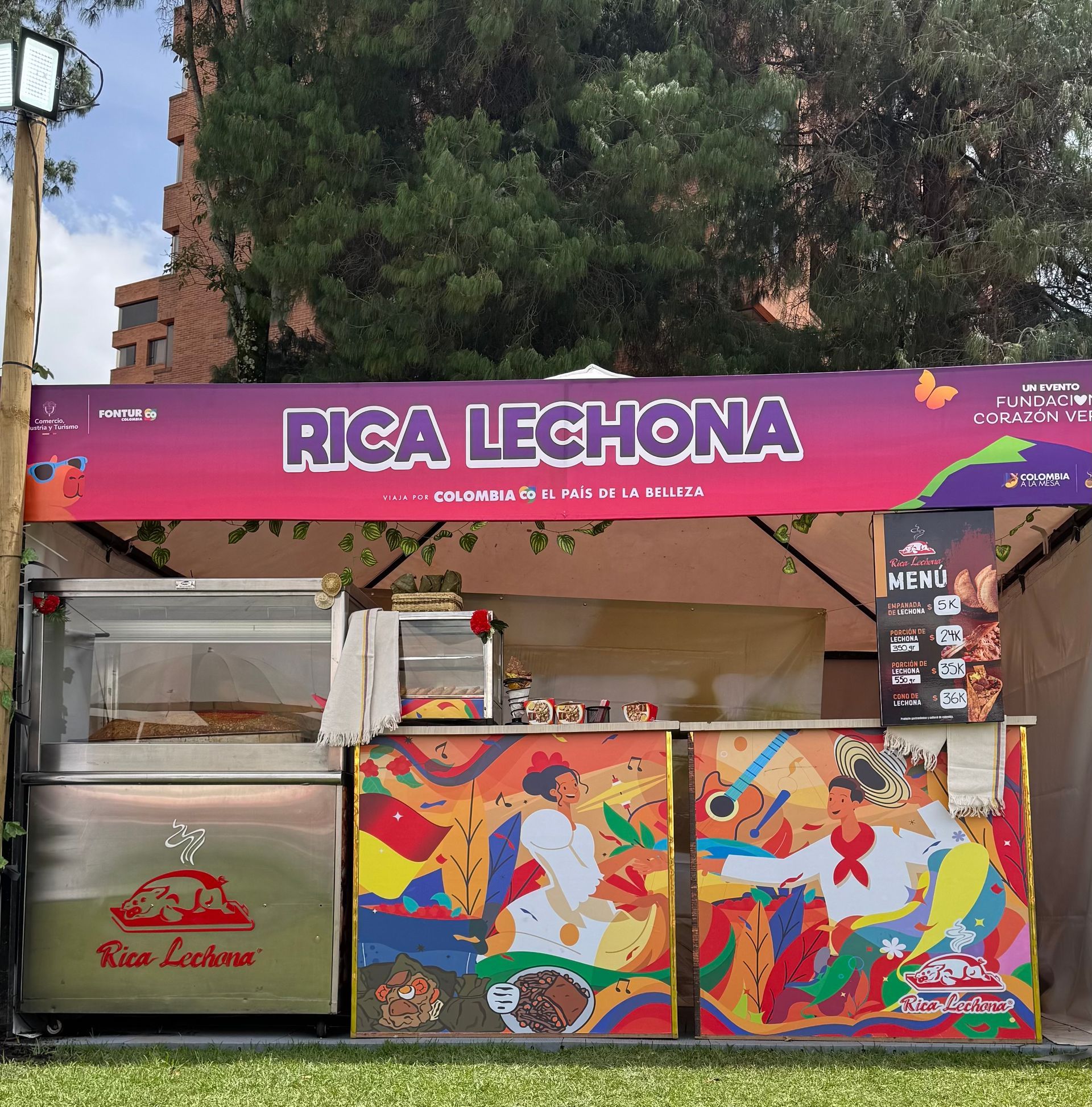 Puesto de comida callejera con la etiqueta “Rica Lechona”, un letrero rosa brillante y coloridos paneles murales.