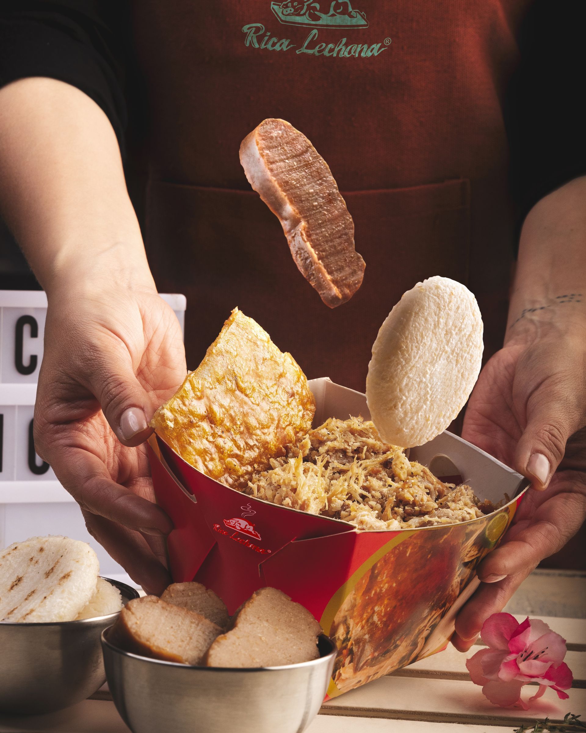 Manos que sostienen un tazón rojo con una variedad de bocadillos fritos, con pan y pasteles flotando sobre él.