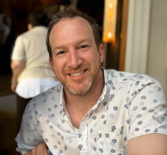 Smiling man in a white patterned shirt seated indoors at a restaurant table