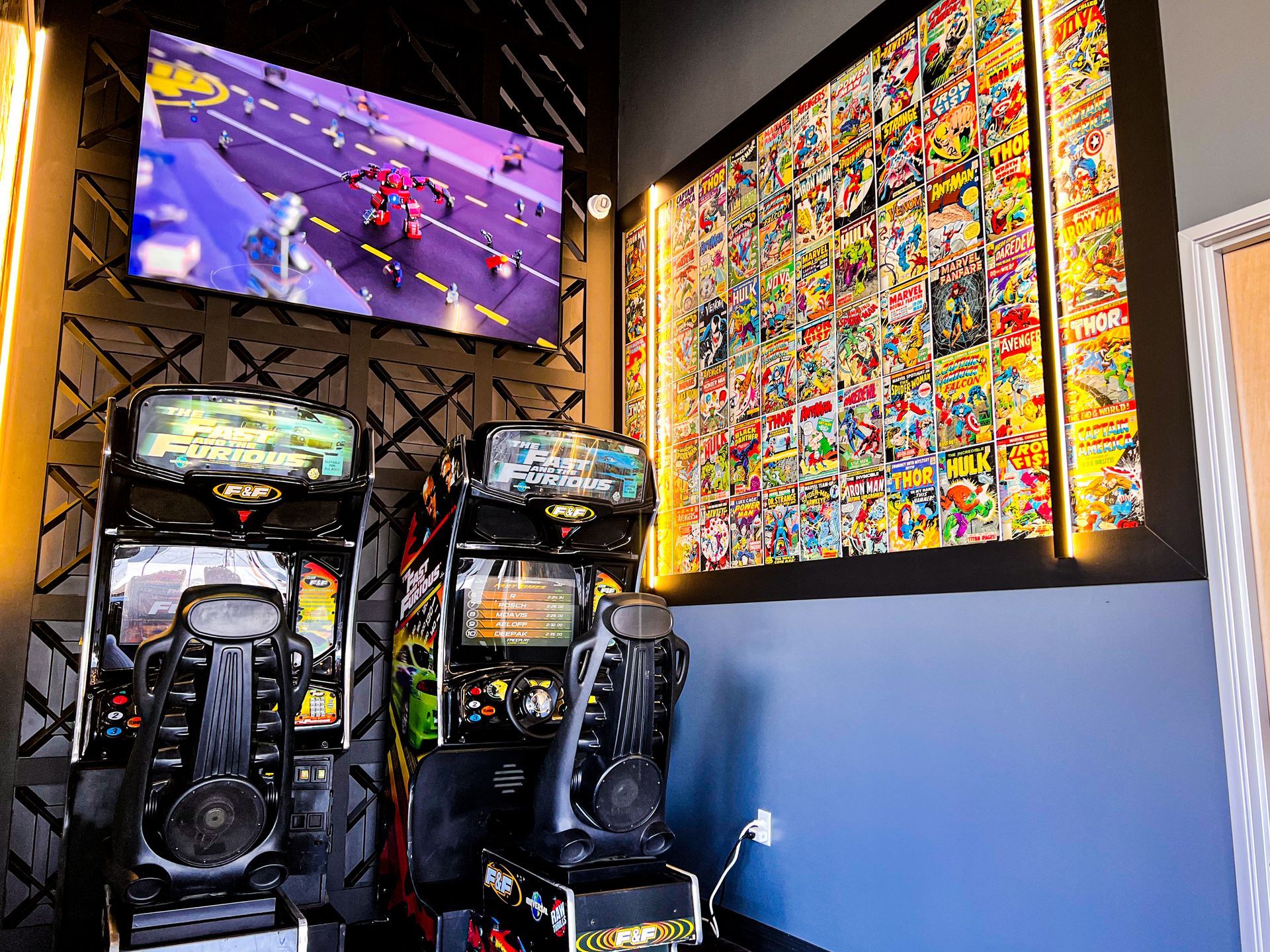 Arcade with two racing game cabinets, a wall mural, and a large TV showing a motorcycle race.