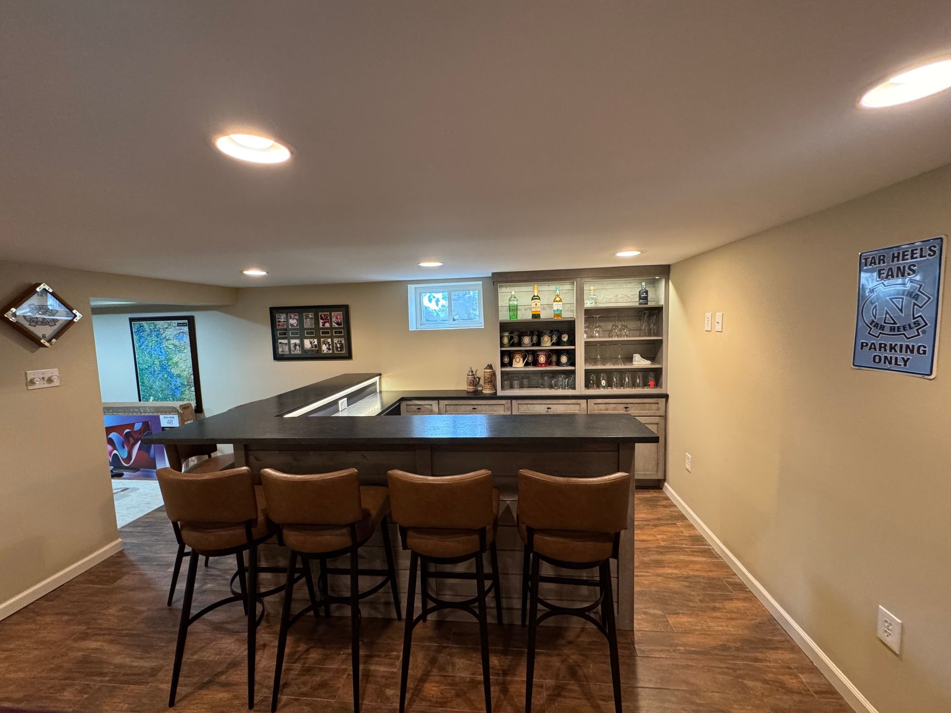 A basement bar area with four brown leather stools, a dark countertop, and shelving stocked with glasses and bottles.