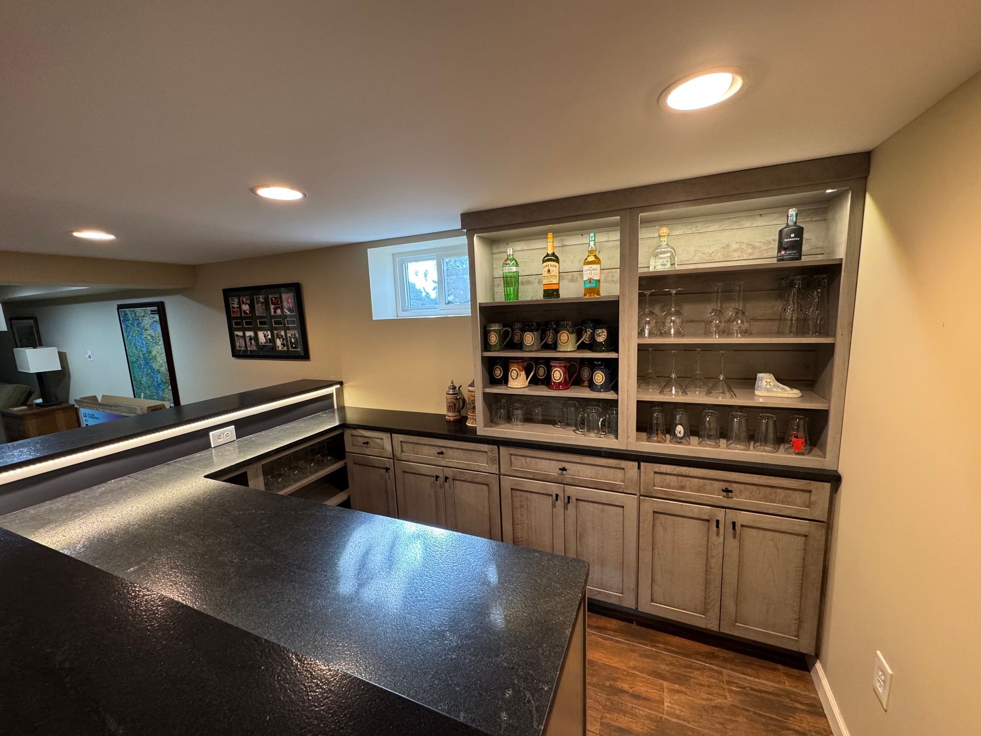 A home bar with gray wood cabinets, black countertops, and built-in shelving displaying glassware and liquor bottles.