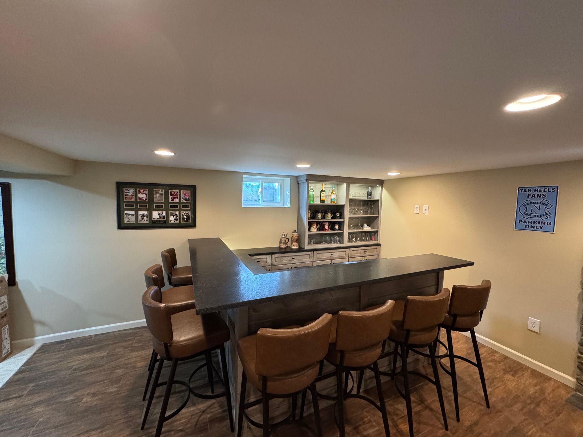 A home basement bar with a dark L-shaped counter and five brown leather stools on a wood-look floor.
