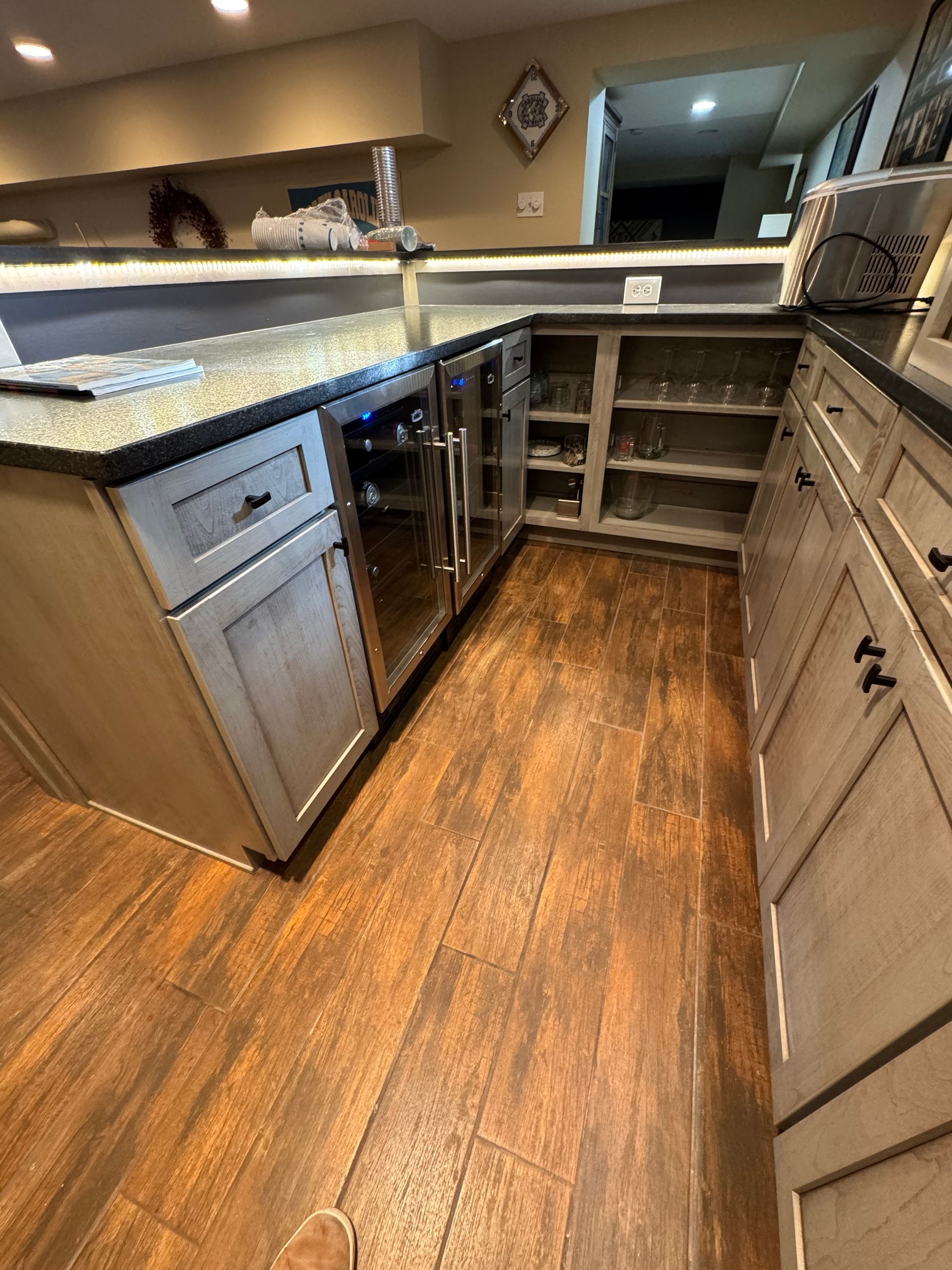 A home bar area with light-toned wooden cabinets, dark countertops, a built-in wine fridge, and wood-look flooring.