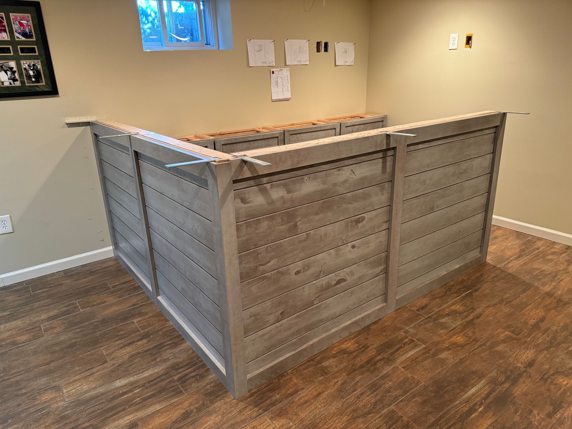 An L-shaped bar with horizontal, light gray wood-look paneling sits in a basement with dark wood flooring.