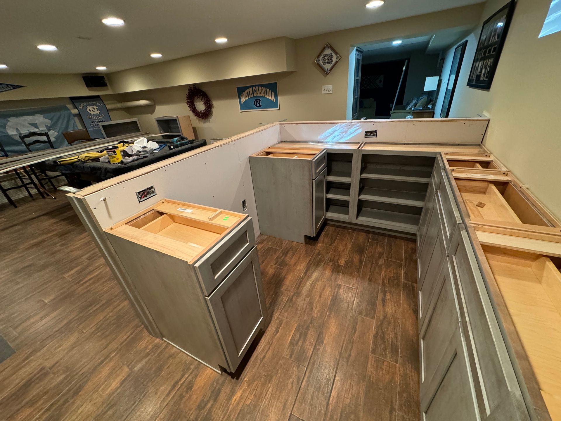 Grey kitchen cabinets being installed in a room with wood-look flooring and a partial wall, during a renovation project.
