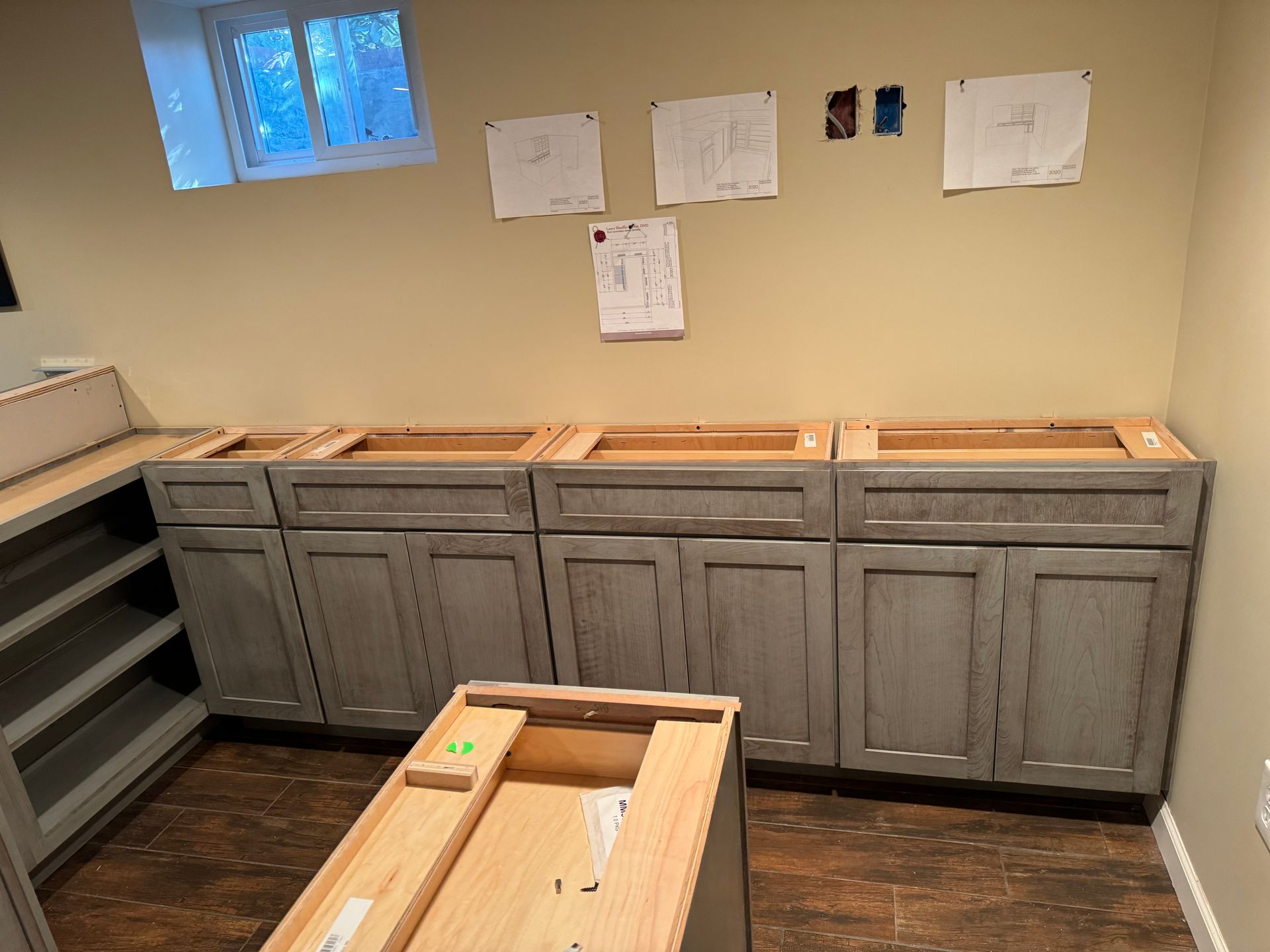Three grey kitchen base cabinets sit on a wood floor in an unfinished room with papers taped to the wall.