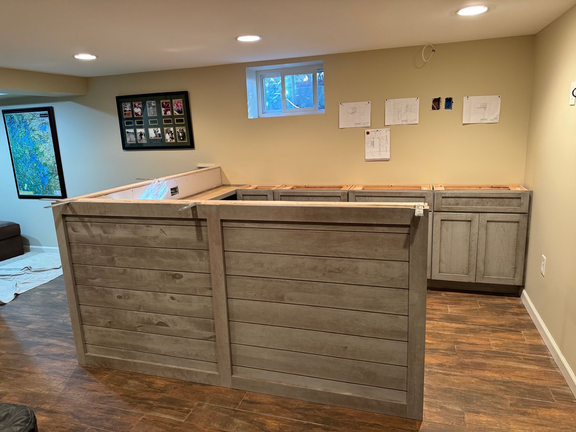 An L-shaped wooden bar structure under construction in a basement, with unfinished base cabinets and a small window.