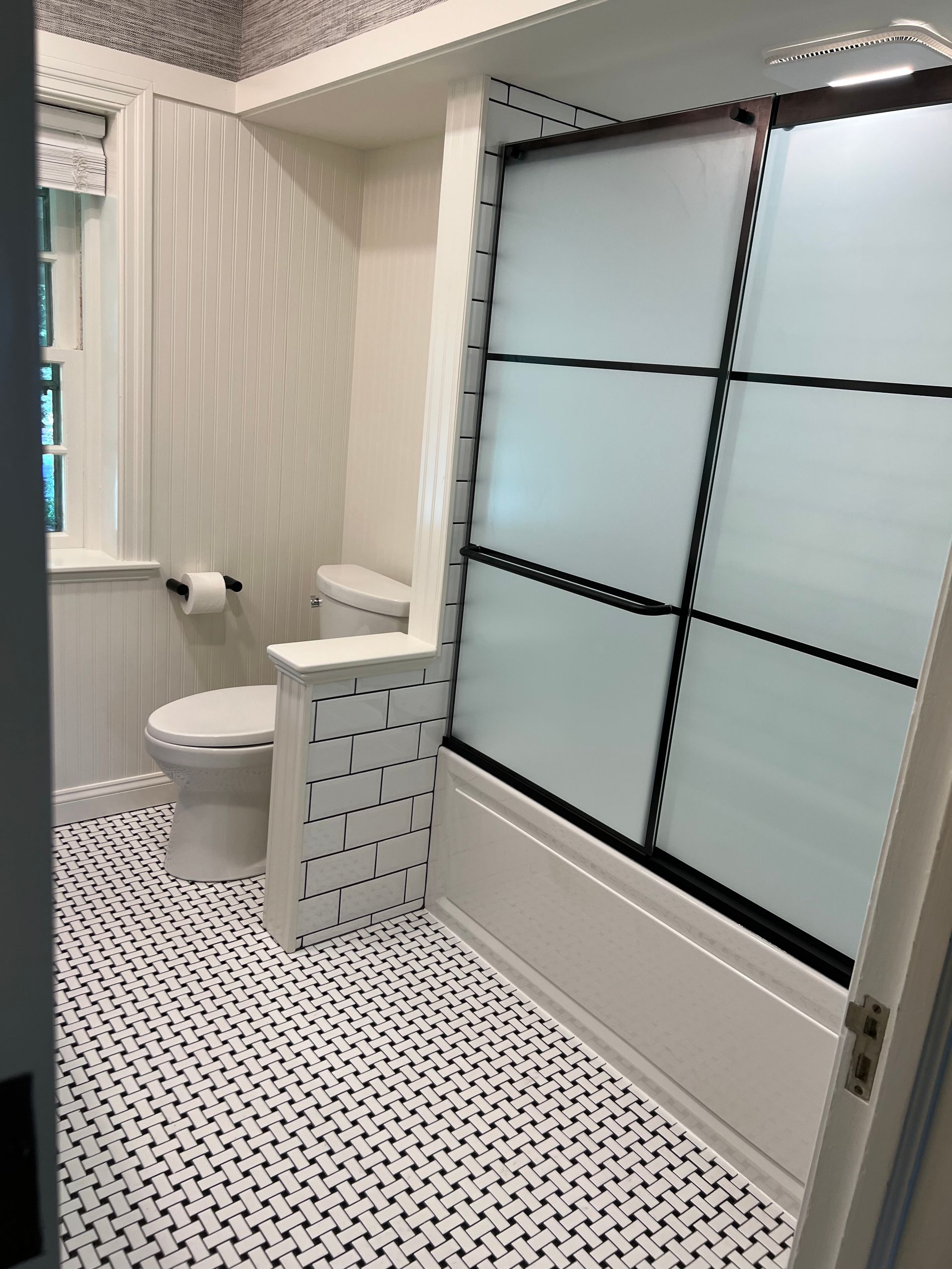 A bathroom with a black-framed, frosted glass shower door, a white toilet, and patterned black and white floor tiles.