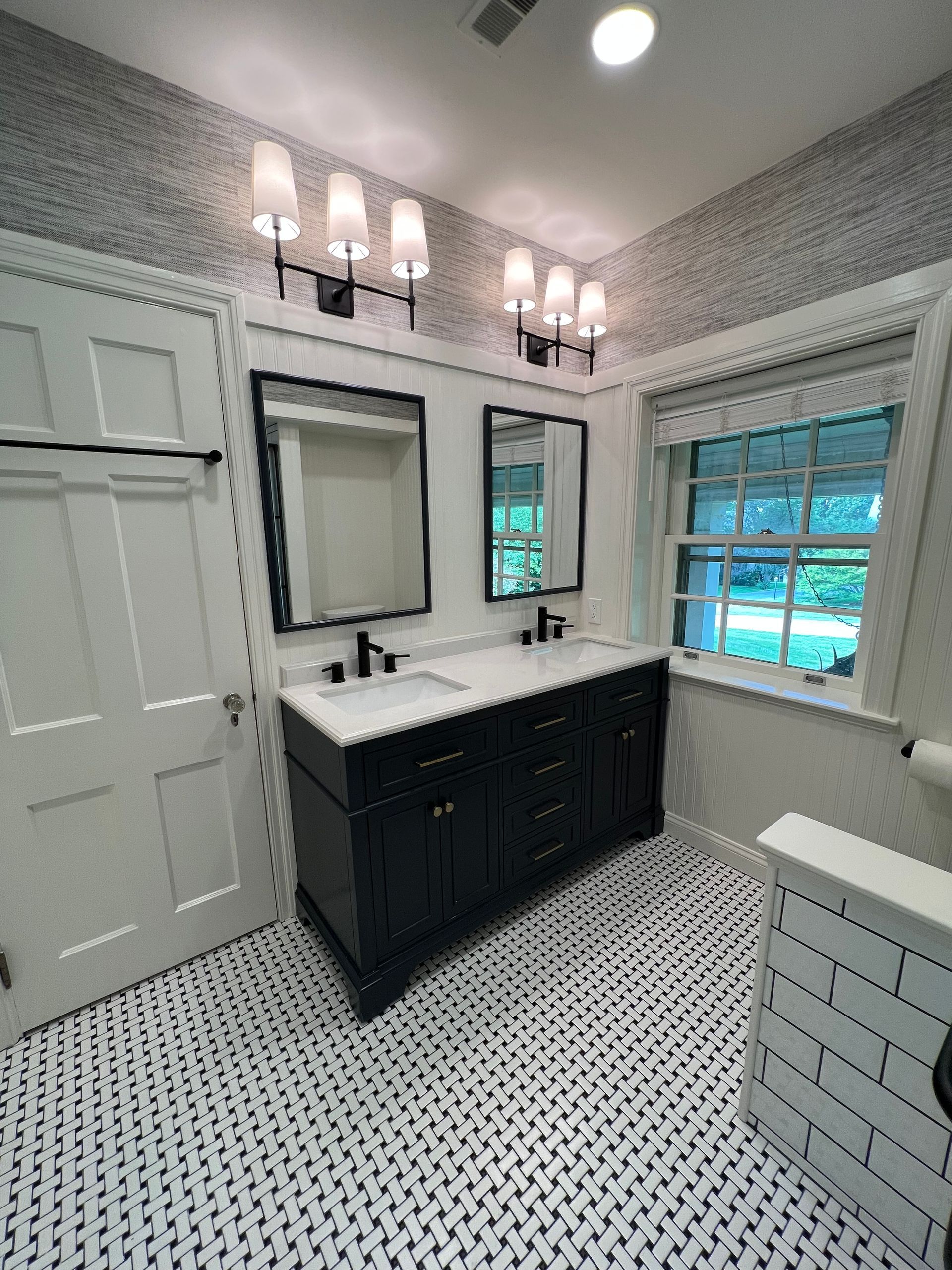 A bathroom with a dark double vanity, two mirrors, patterned floor tile, and white subway tile wainscoting.