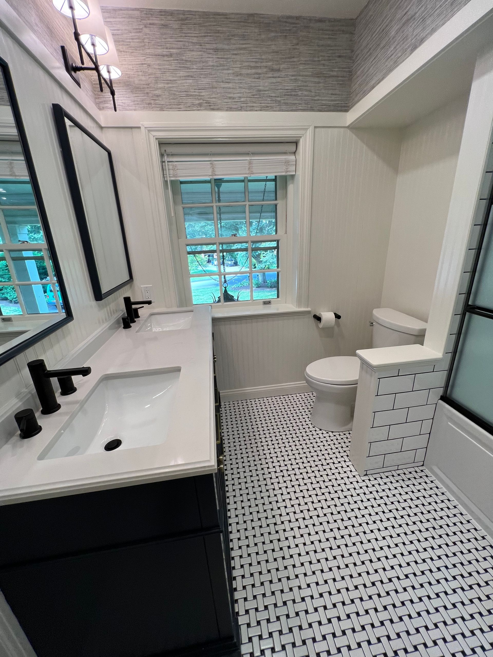 A bathroom featuring a double vanity with black faucets, patterned floor tiles, a window, and a white toilet.